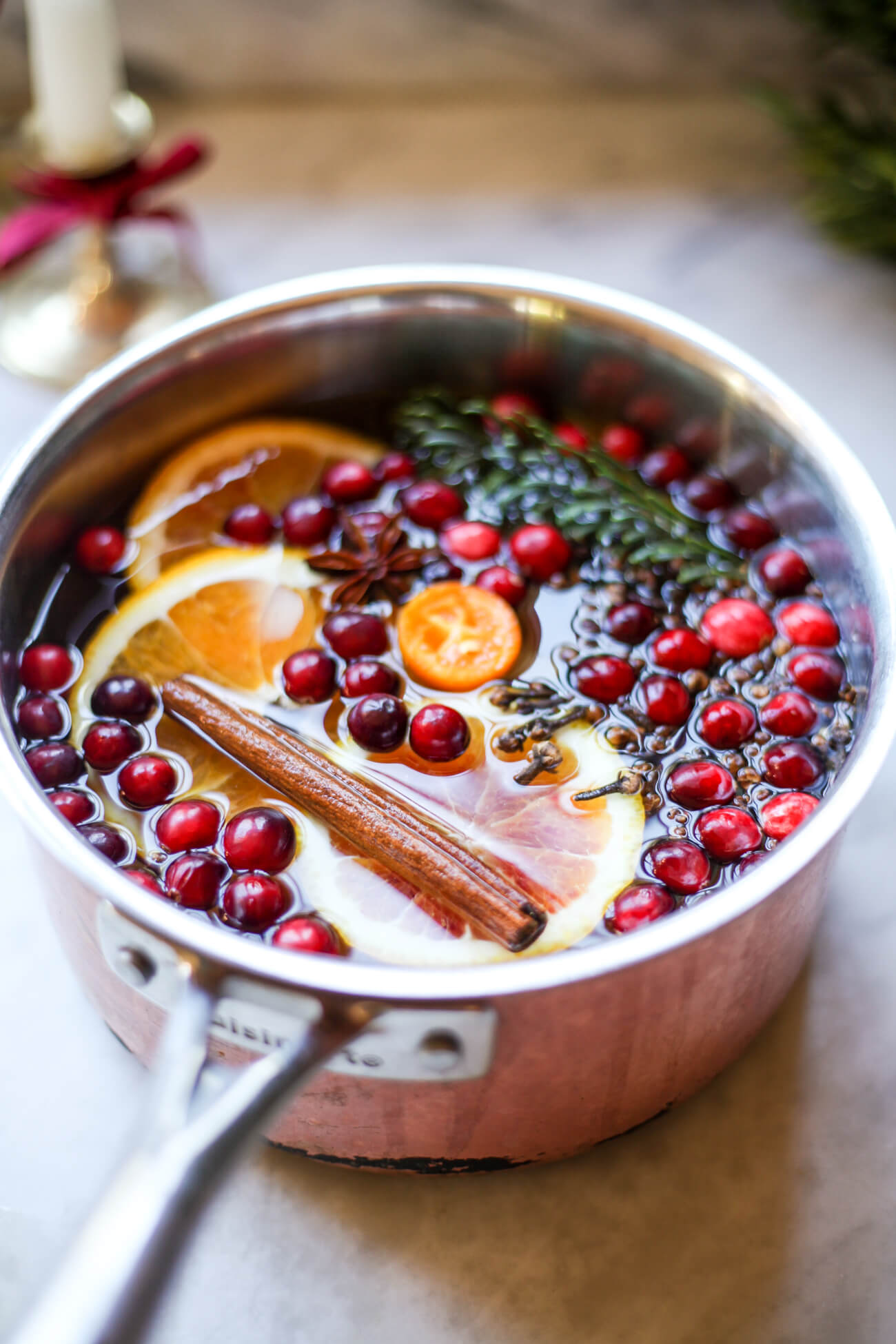 A medium-sized copper saucepan filled with a Christmas stovetop potpourri recipe. Fresh cranberries float in water with orange, a kumquat, cinnamon sticks, cloves, star anise, and pine. 