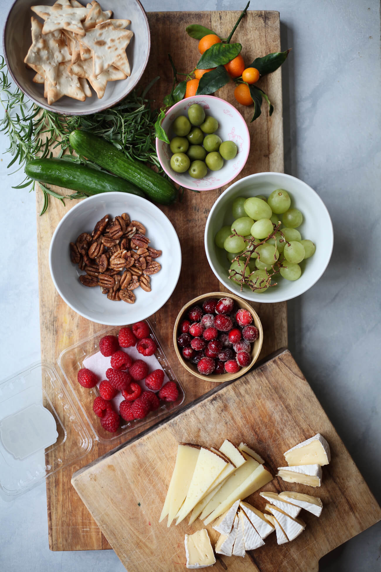 Assorted ingredients for a Christmas tree charcuterie board, including star crackers, pecans, brie, manchego, grapes, olives, cucumbers, kumquats, raspberries, cranberries, and rosemary on wooden boards.
