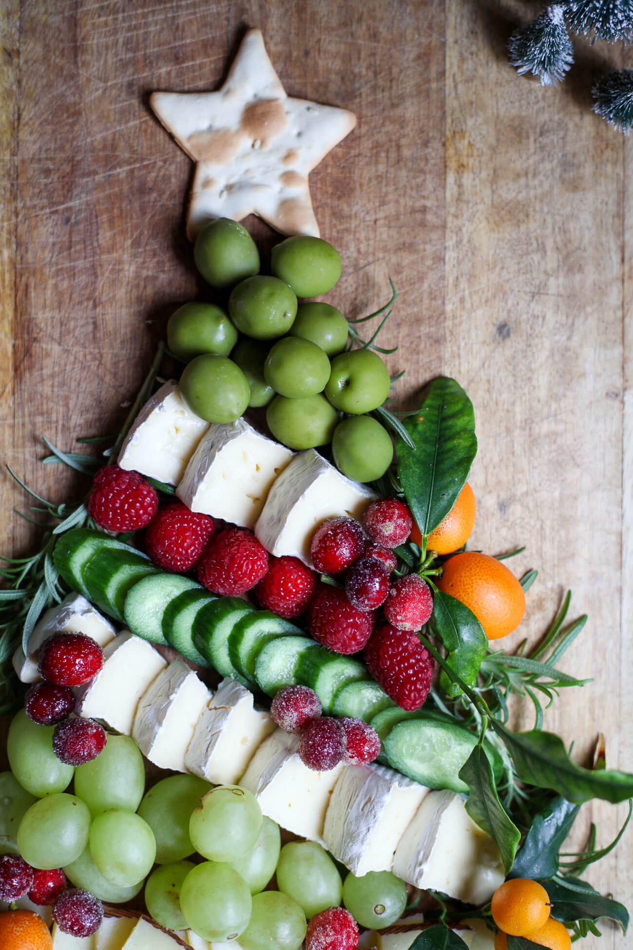 Top section of a Christmas tree charcuterie board featuring a star-shaped cracker, green grapes, brie cheese, raspberries, cucumbers, kumquats, and rosemary garnish.