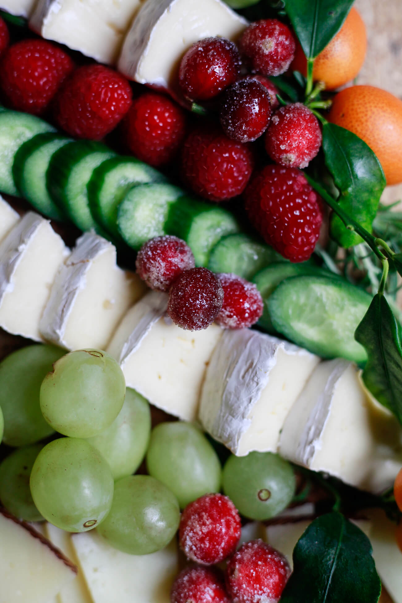 Close-up view of brie cheese slices, green grapes, cucumbers, raspberries, sugared cranberries, and kumquats arranged for a festive Christmas tree charcuterie board.
