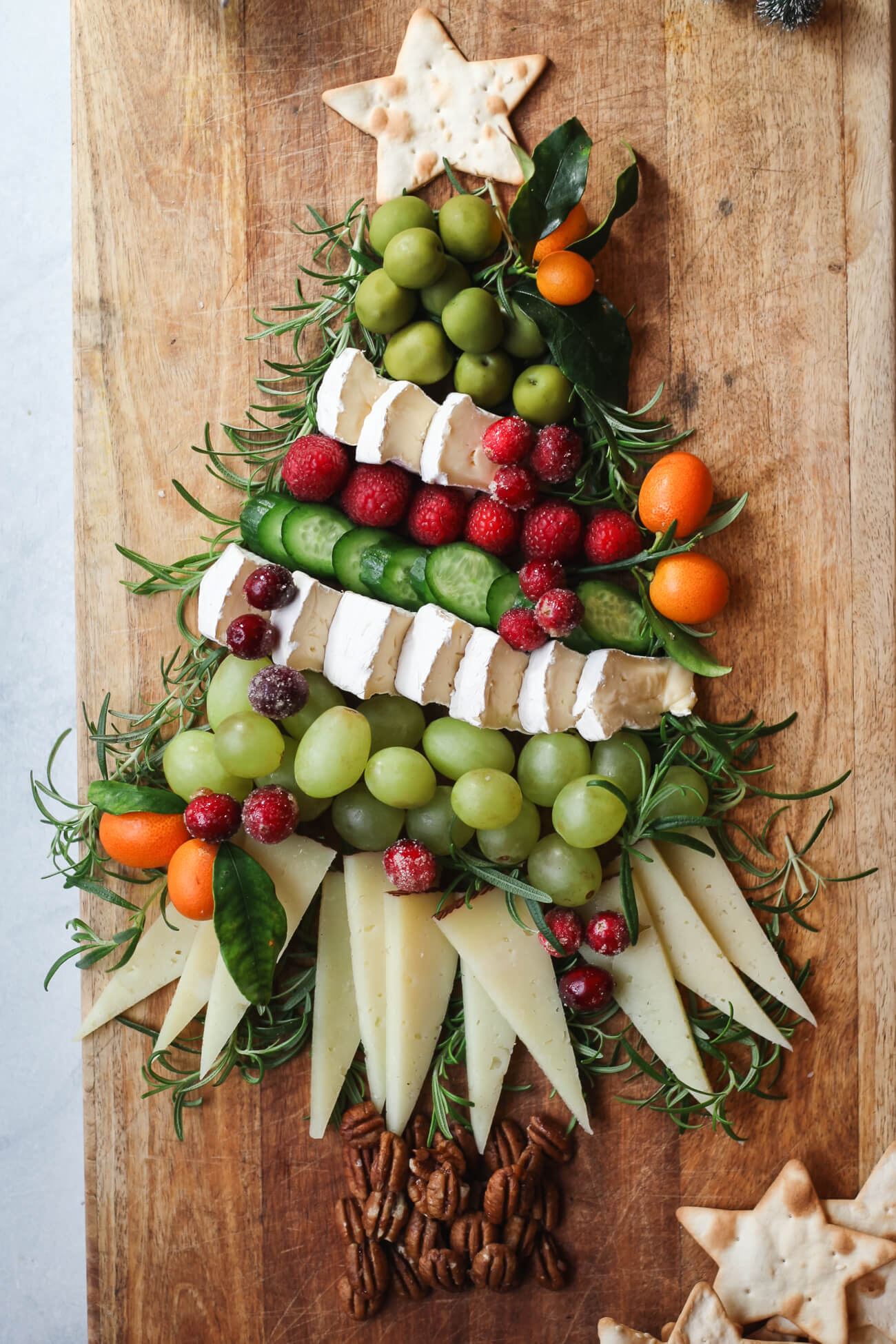 Festive charcuterie board arranged in the shape of a Christmas tree with brie, manchego, grapes, cucumbers, raspberries, kumquats, pecans, rosemary, and a star-shaped cracker topper.
