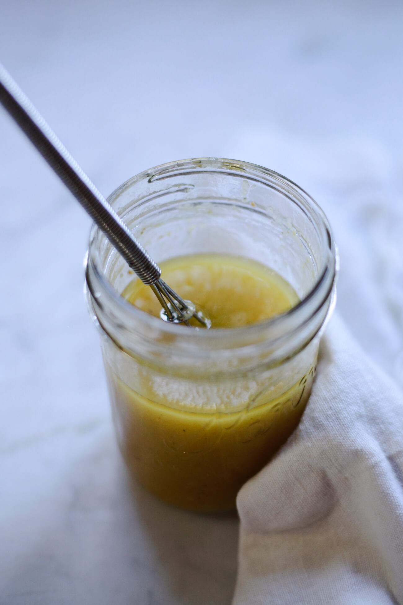 A small jar filled with a homemade lemon Dijon vinaigrette with a small whisk.