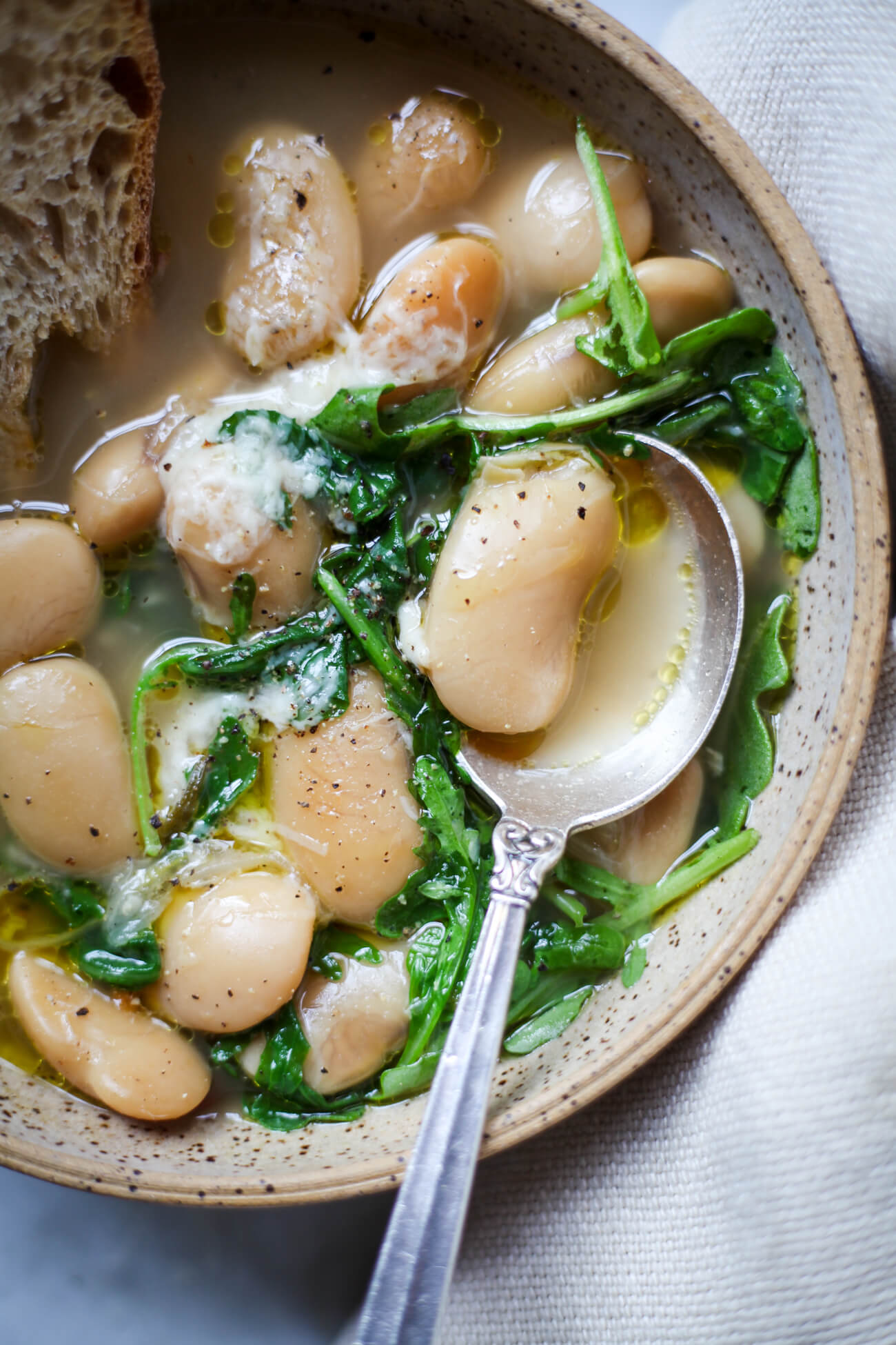A close-up photo of a giant white Royal Corona beans in broth with wilted arugula in a ceramic bowl with a silver soup spoon. A perfect bowl of brothy beans. 