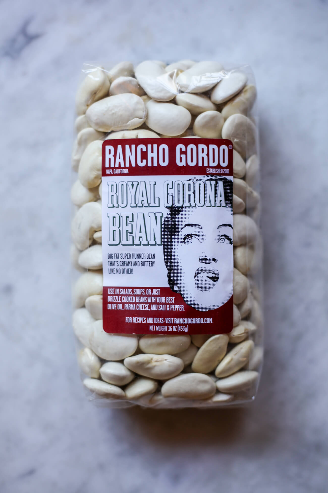 A bag of Rancho Gordo Royal Corona Beans in their original packaging on a white marble countertop. This is a bag of giant white beans. 