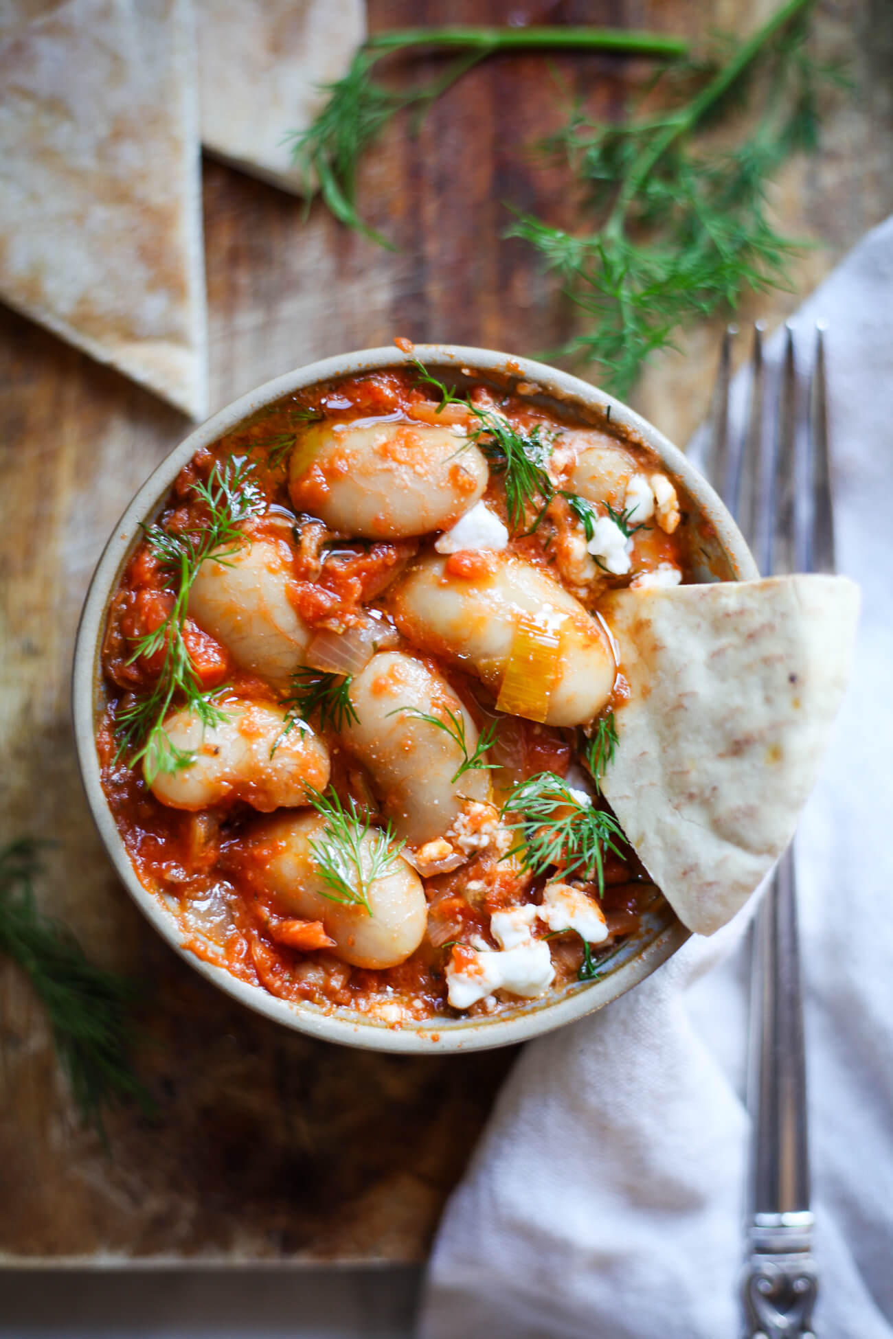 An overhead photo of a bowl filled with Greek giant beans in red sauce (Gigantes Plaki) topped with fresh dill and feta and served with pita bread. 