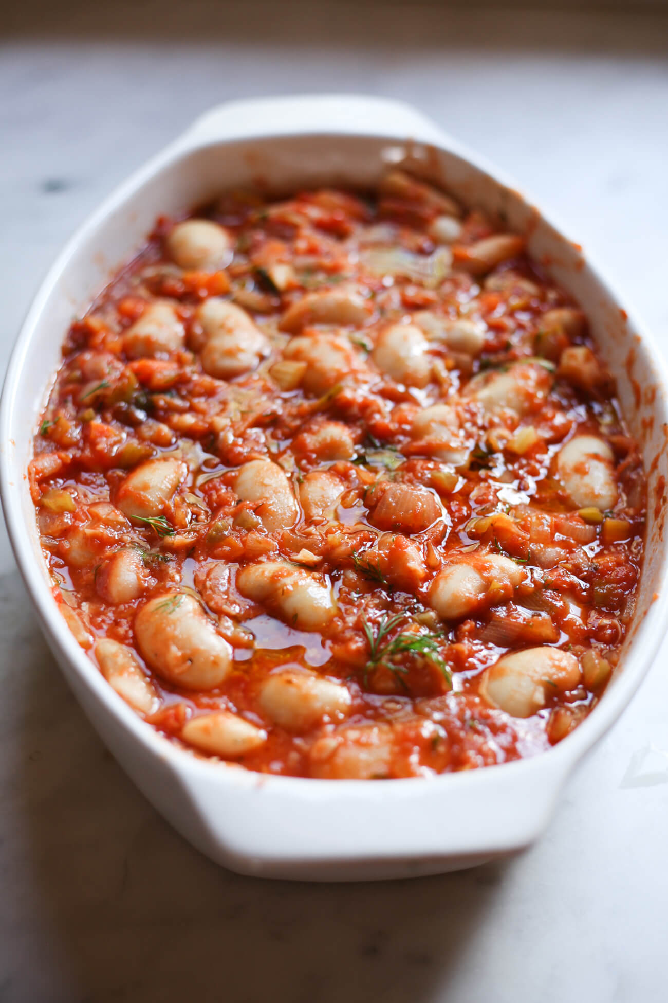 A white oval casserole dish filled with cooked giant Greek white beans and tomato sauce before cooking. This is how to make Gigantes Plaki, a vegetarian Greek recipe.