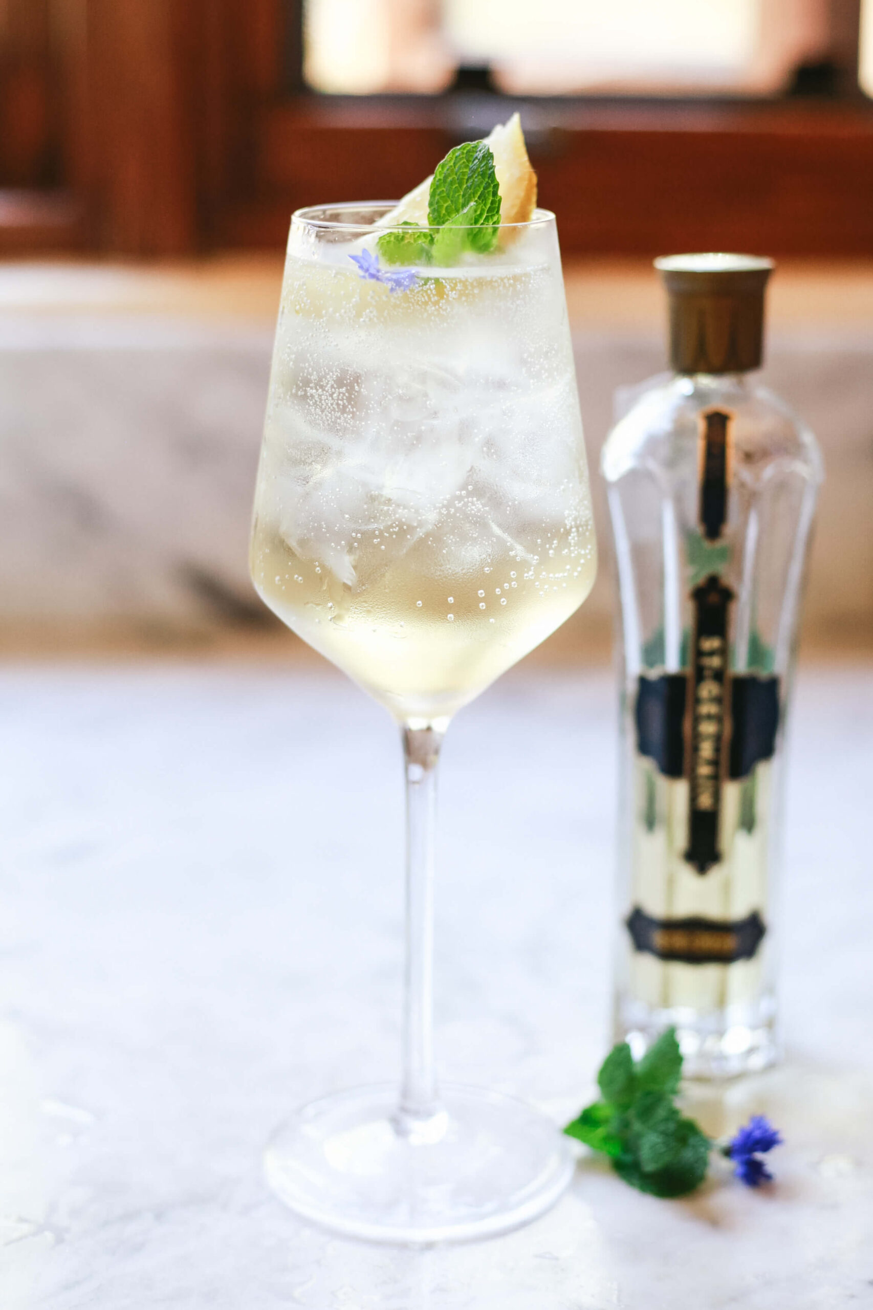 Glass of Hugo Spritz cocktail with ice, mint, and lime garnish, served in a wine glass with a bottle of St-Germain elderflower liqueur in the background.