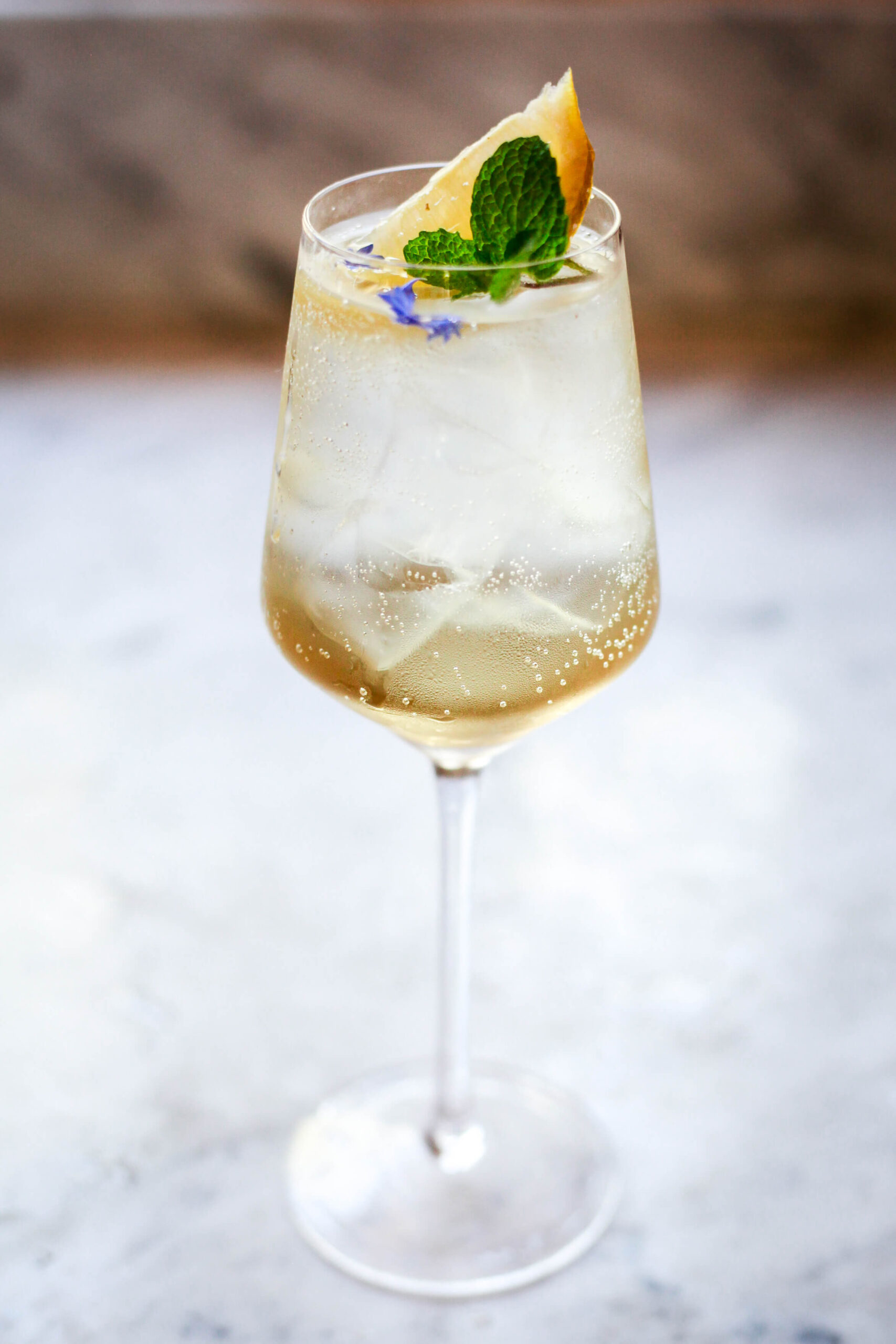 Tall wine glass filled with Hugo Spritz cocktail, garnished with lemon wedge, mint sprig, and purple flowers on ice.