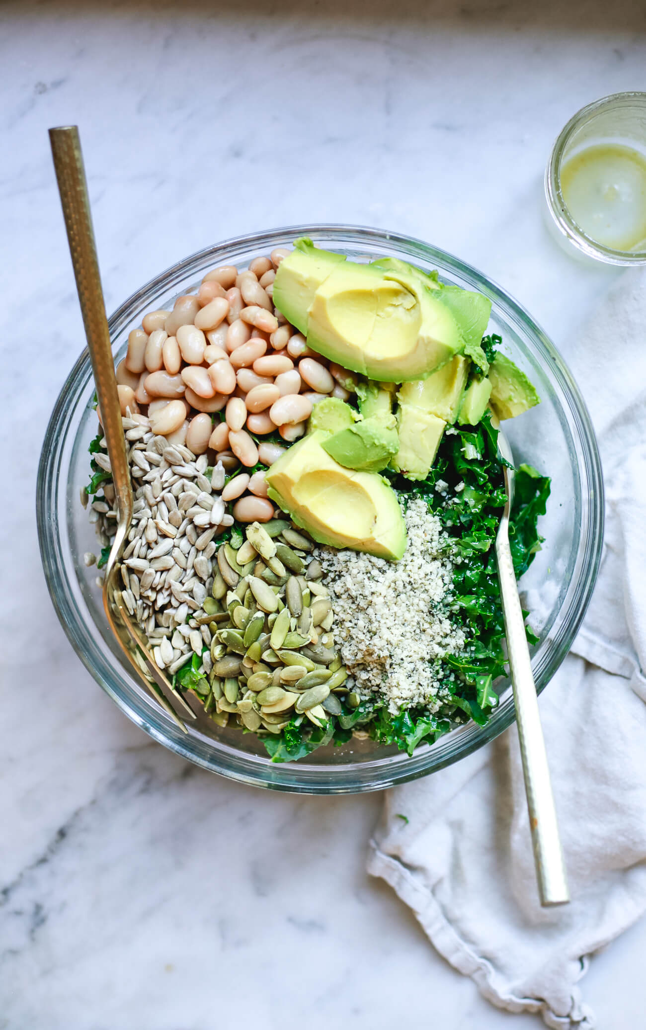 An overhead photo of a glass bowl filled with shredded kale, pumpkin seeds, sunflower seeds, hemp seeds, white beans, and avocado on a marble countertop. 