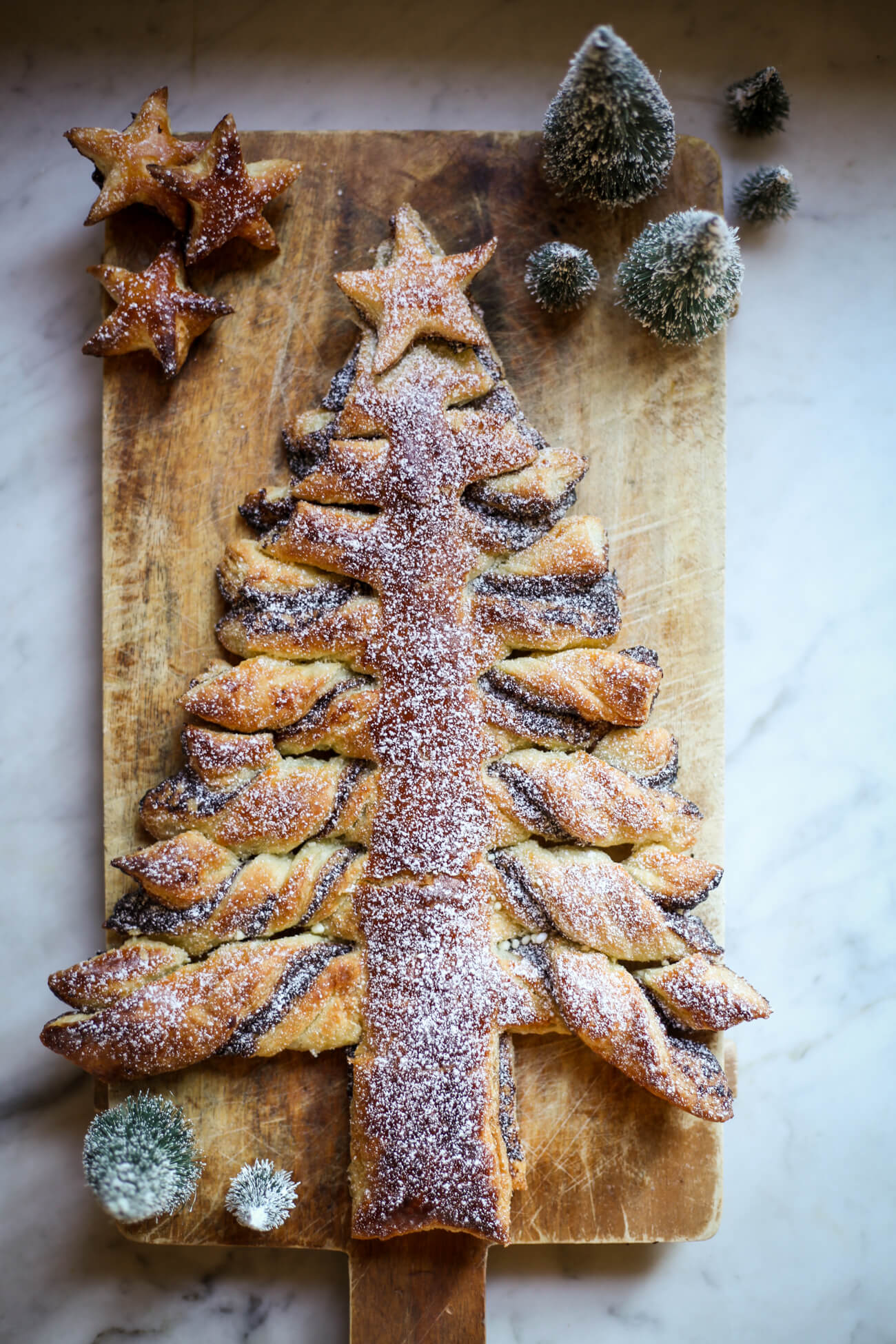A puff pastry nutella Christmas tree dusted with powdered sugar on a cutting board decorated with faux Christmas tres and puff pastry stars.