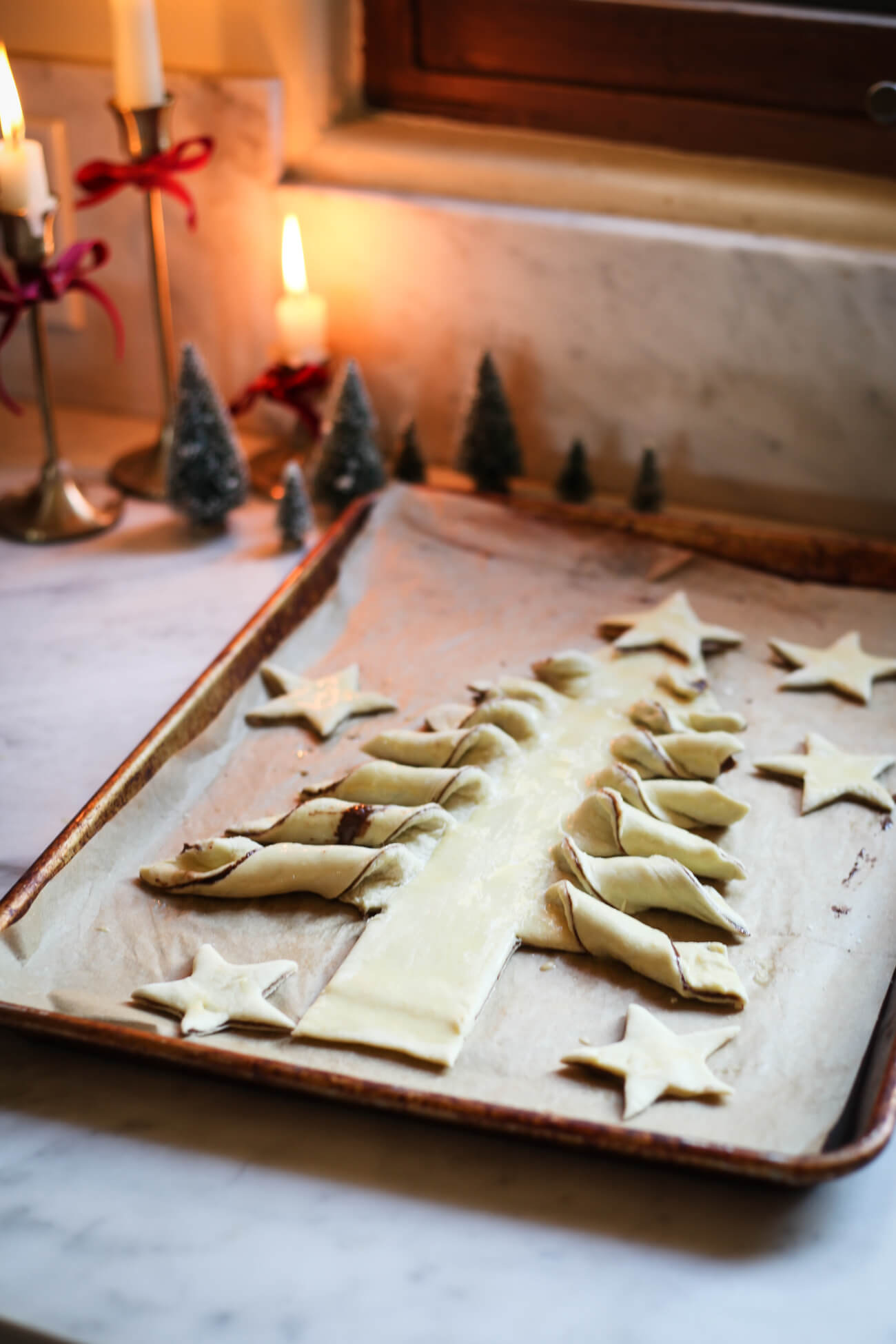 A Nutella puff pastry Christmas tree on a baking sheet in a marble kitchen before going into the oven. The Christmas tree branches are twists. Cut out puff pastry stars are around the tree. 