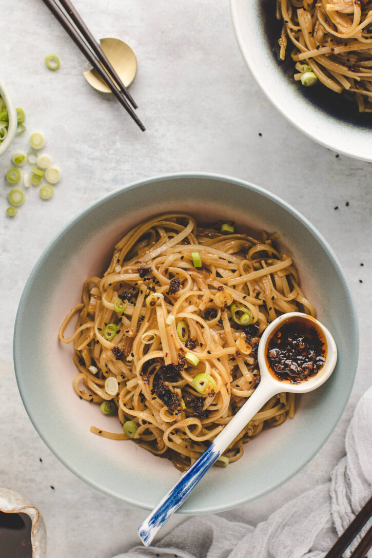 Bowl of spicy chili garlic noodles topped with green onions and served with a spoonful of chili oil on the side.