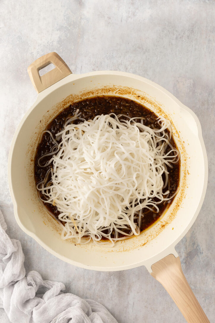 Cooked rice noodles added to a skillet with rich chili garlic sauce, ready to be tossed together for chili garlic noodles.