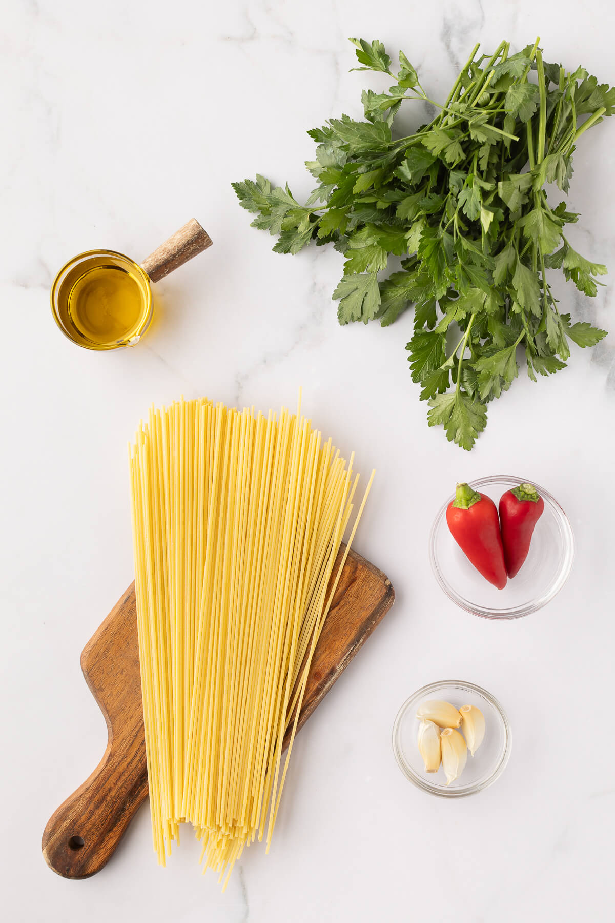Olive oil, fresh Italian parsley, Italian peperoncino, garlic cloves, and spaghetti sit on a marble countertop. 