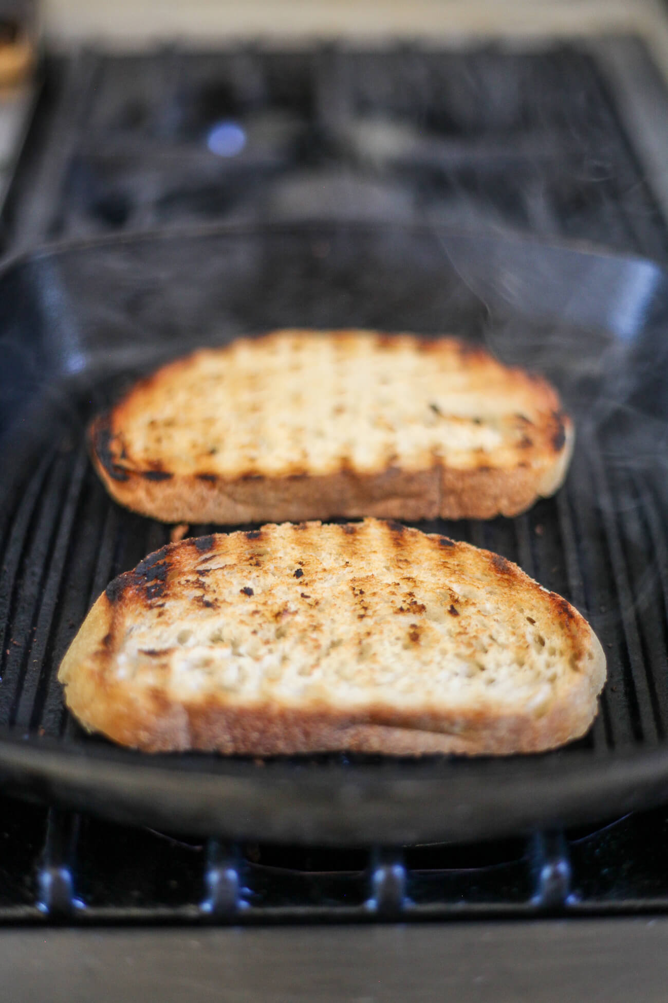 Two pieces of Tuscan bread with grill marks continue to cook on a black grill pan to make fettunta bruschetta.