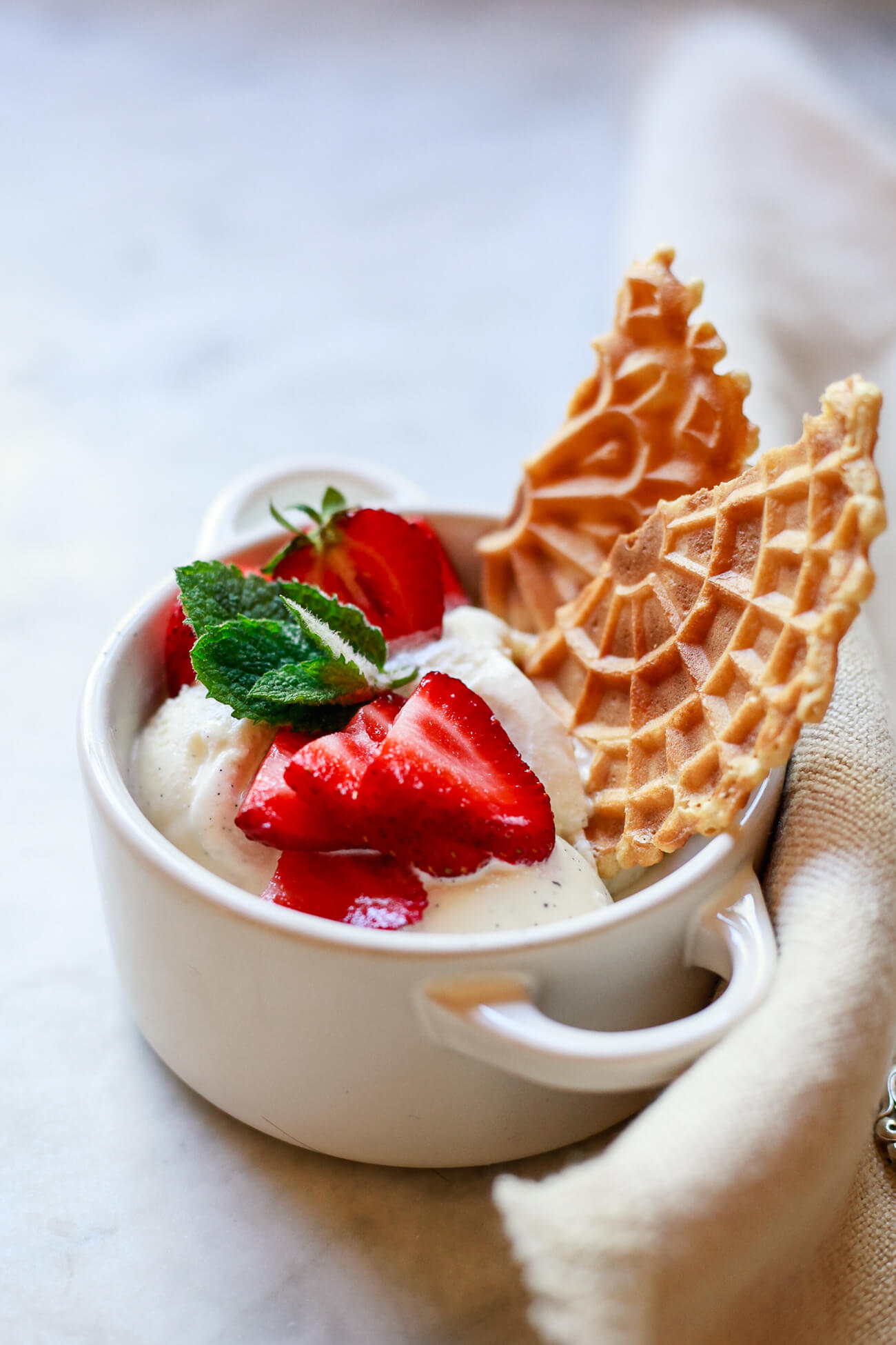 A white ceramic ramekin filled with vanilla bean ice cream topped with sliced strawberries, a sprig of mint, and a pizzelle cookie. An easy Italian dinner party dessert. 