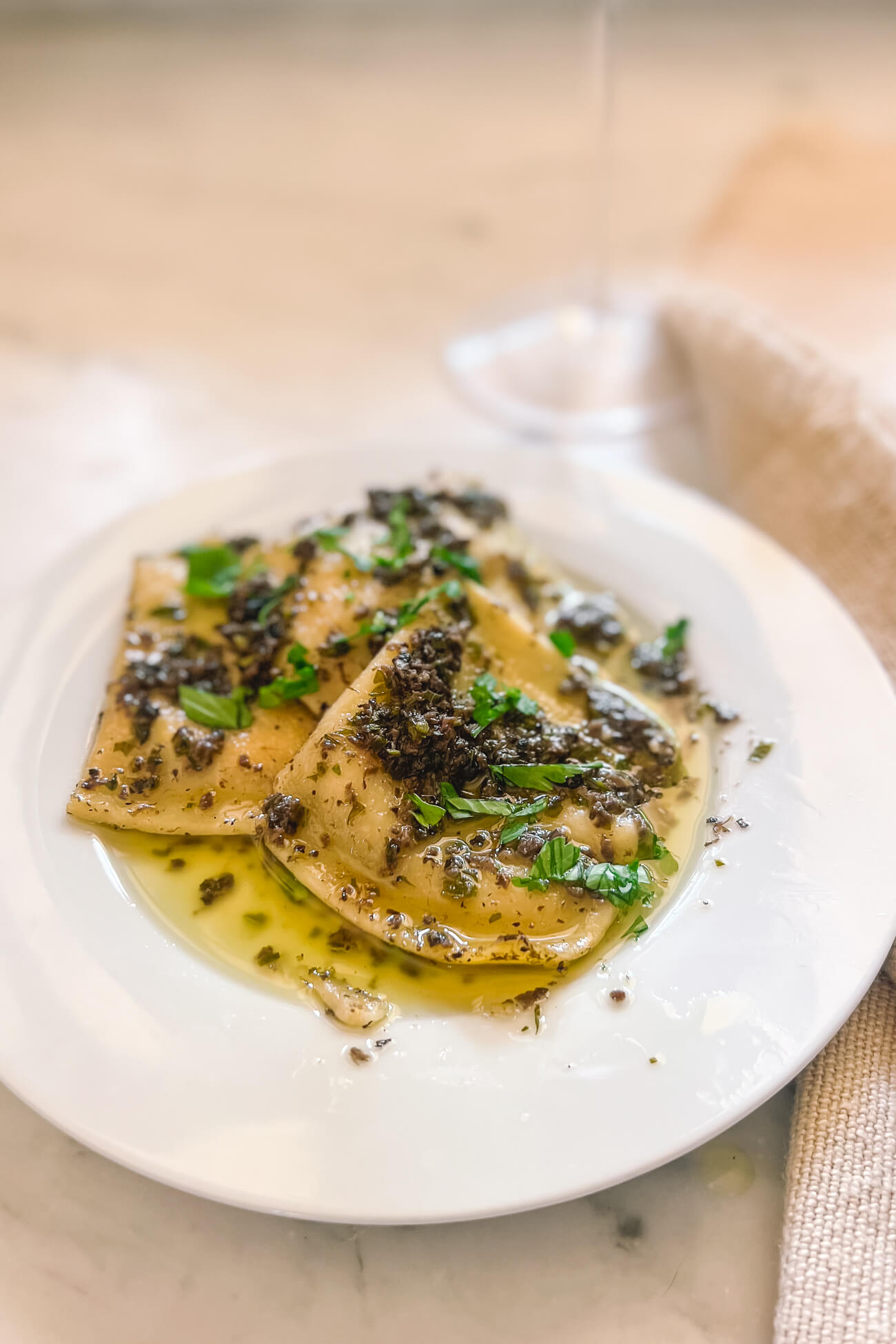 A white plate with ravioli covered in black truffle sauce garnished with parsley. 