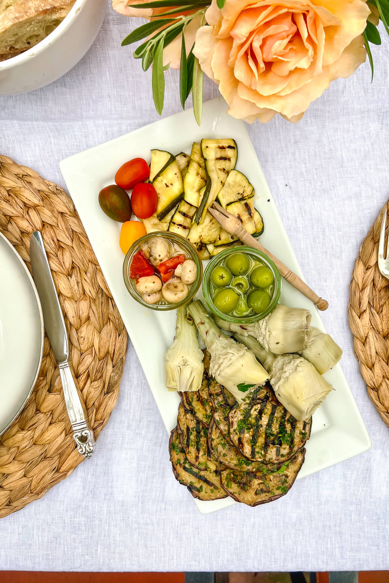 A white rectangular platter topped with antipasti: grilled zucchini, grilled eggplant, artichokes, olives, marinated mushroooms, and cherry tomatoes. 