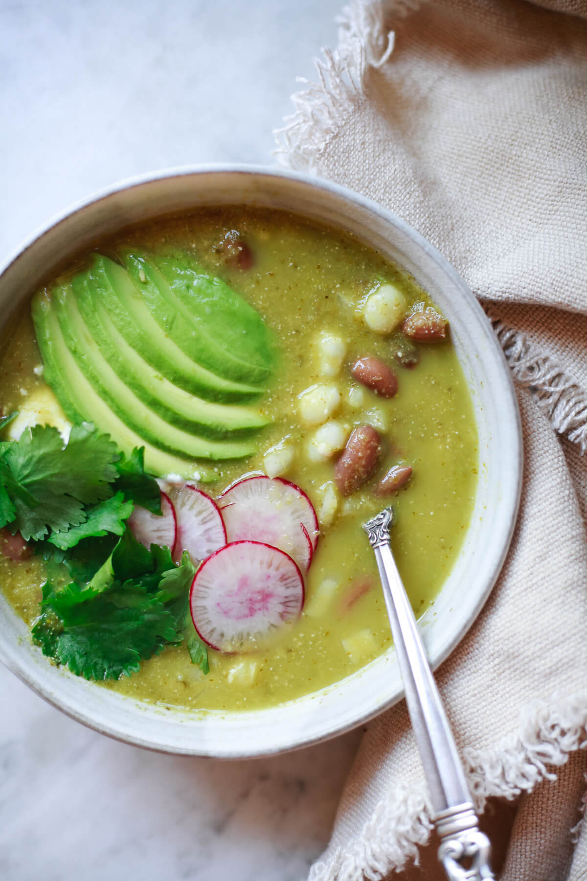 An overhead photo of a grey ceramic bowl filled with pozole verde with white hominy and pinto beans. The Mexican soup is topped with sliced avocado, radish, and cilantro and sits on a marble counter. 