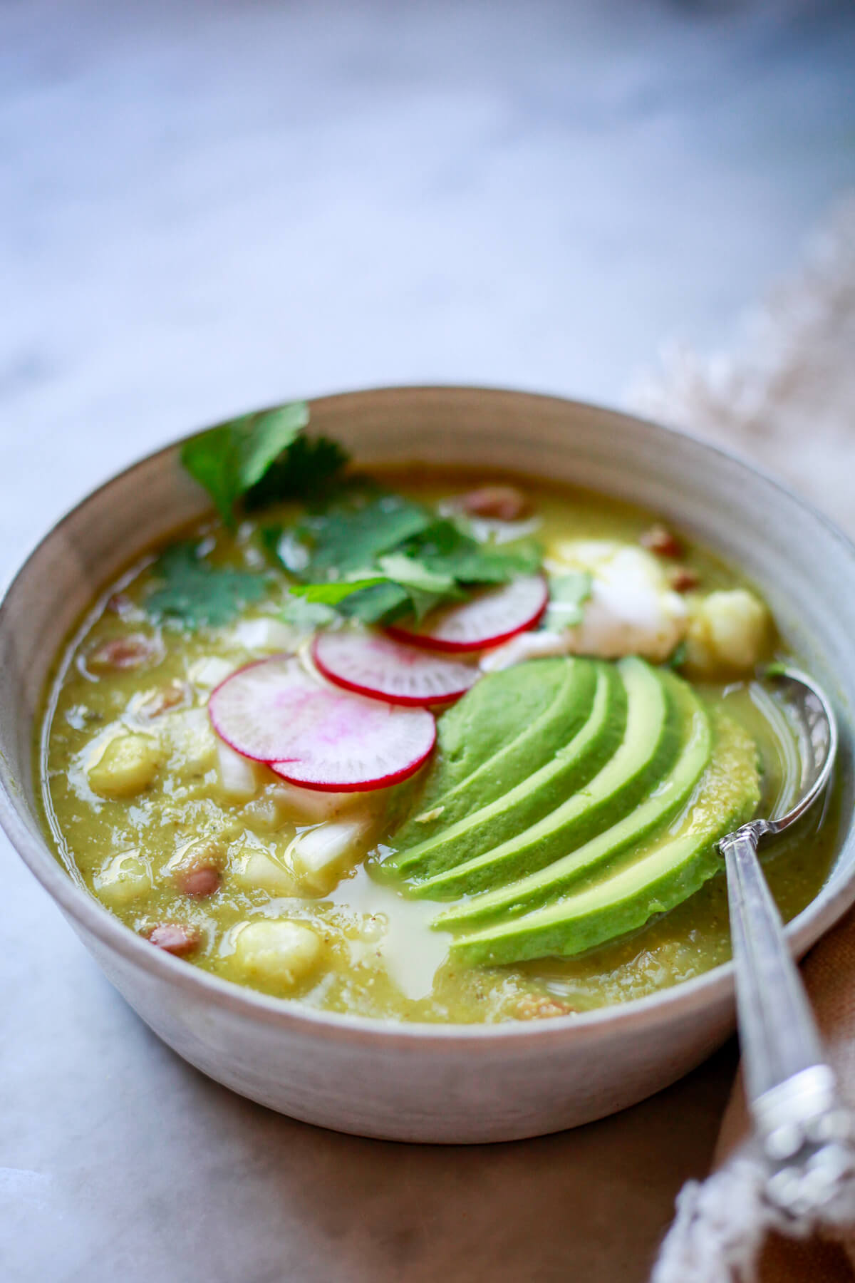 A bowl of vegan pozole in a grey ceramic bowl. A Mexican pinto bean and hominy soup topped with avocado, radish slices, and cilantro. 