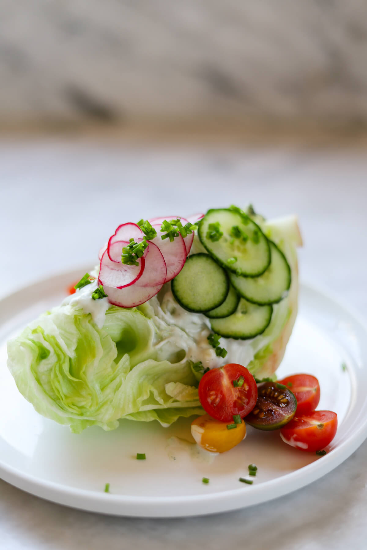 A wedge salad topped with cucumber, radish, tomatoes and chives on a white plate.