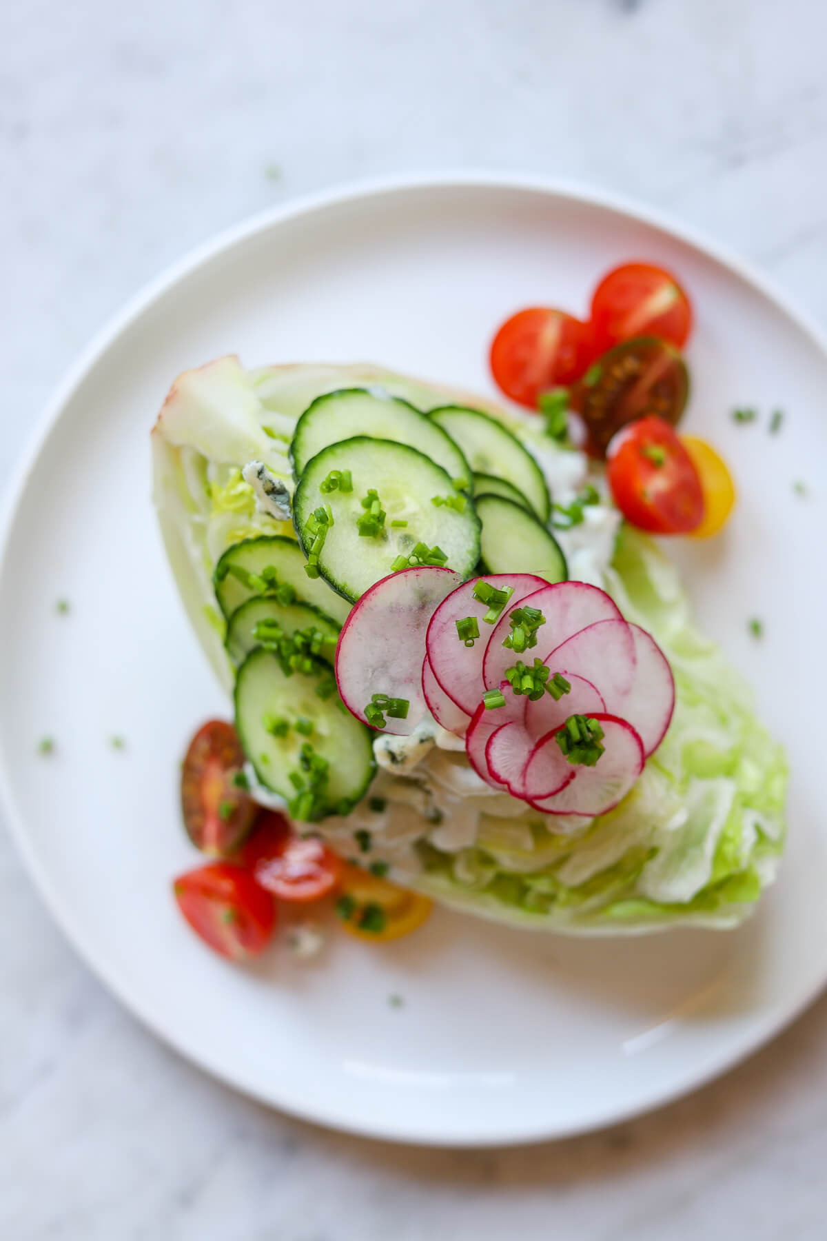 Overhead view of iceberg wedge salad topped with blue cheese dressing, sliced cucumbers, radishes, cherry tomatoes, and chopped chives on a white plate.