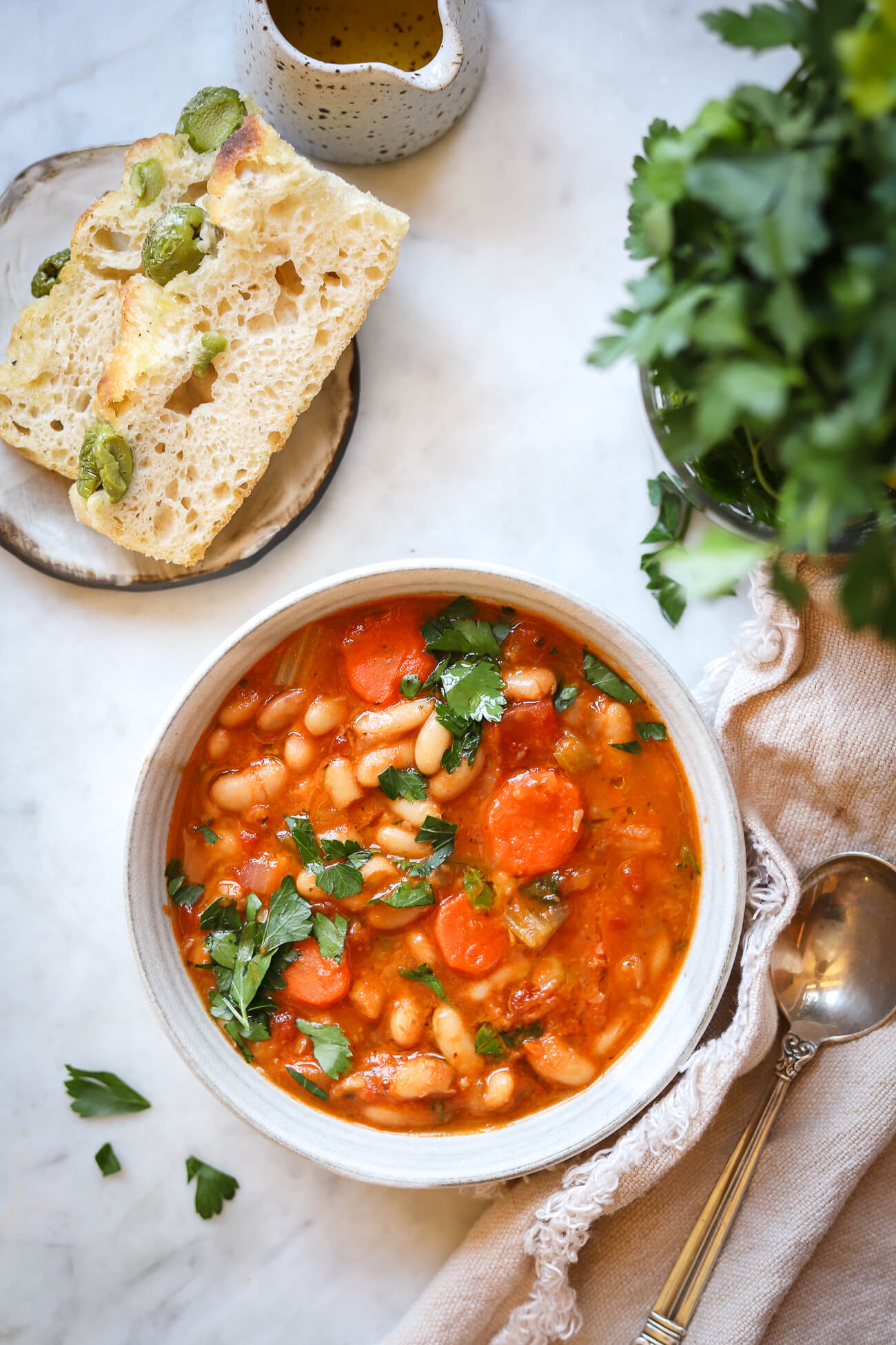 An overhead food photo of Greek white bean soup with carrots and parsley in a light grey ceramic bowl. Olive focaccia and parsley are on the side. 