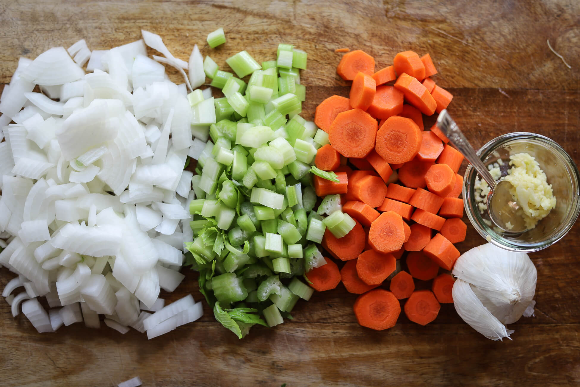 Chopped wh ite onion, celery, sliced carrots, and crushed garlic on a wooden cutting board to prep for Greek bean soup. 
