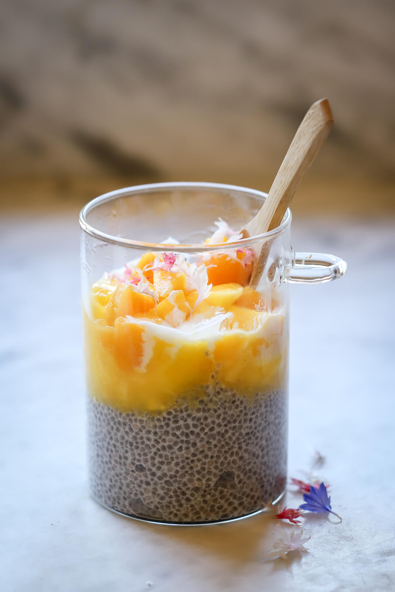 A glass meal-prep overnight oats or chia pudding jar with bamboo spoon filled with chia pudding topped with mango puree, yogurt, mango chunks, and edible flowers on a marble countertop. 