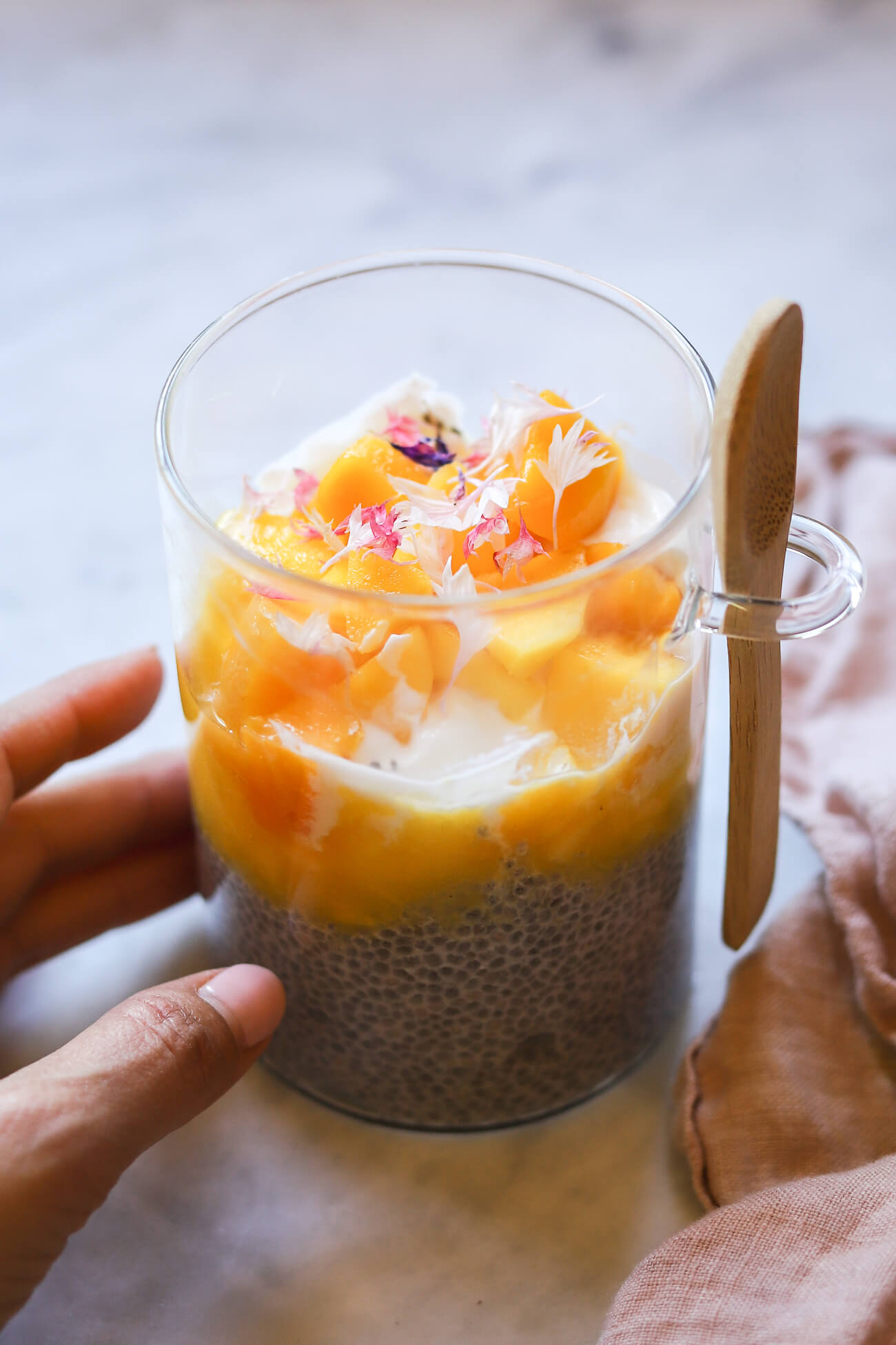A hand reaches in to pick up a glass meal-prep jar filled with delicious mango chia pudding. This is a chia pudding parfait that is layered with chia pudding, yogurt, and mango puree and chunks all topped with edible pink bachelor button flowers. 