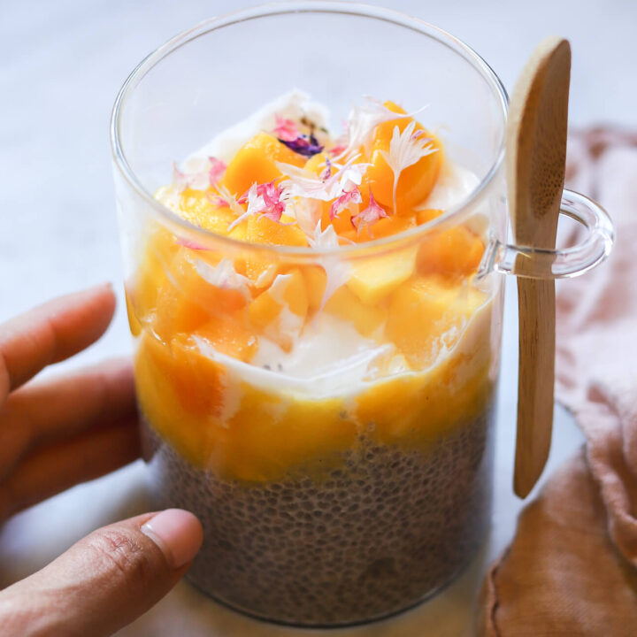 A glass filled with chia pudding topped with yogurt and mango puree and mango chunks with pink edible flowers. The meal-prep glass has a wooden spoon attached.
