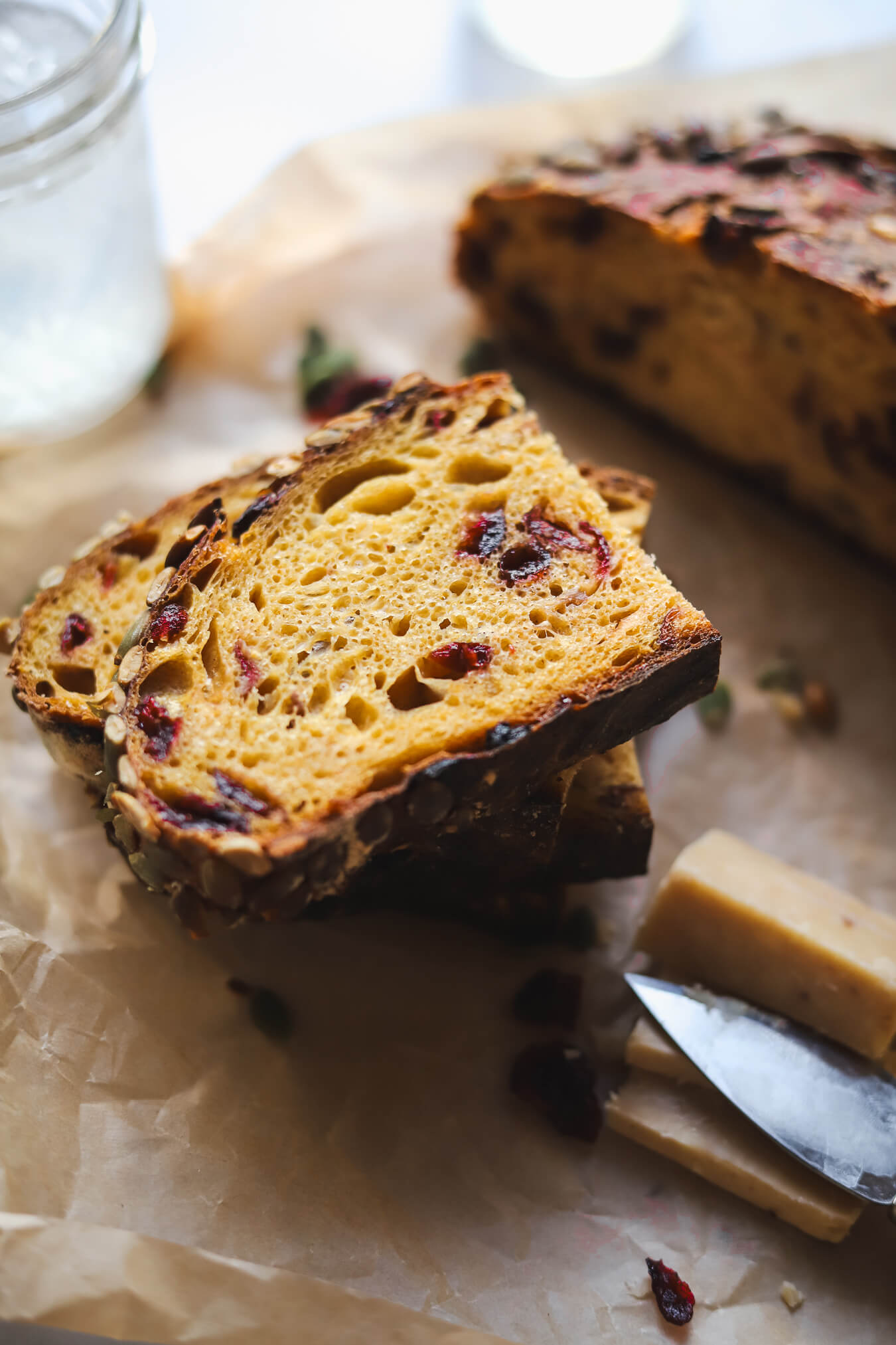 Pumpkin Sourdough Bread with Cranberries Slices of a seeded pumpkin sourdough bread loaf with dried cranberries and pumpkin seeds on a piece of parchment paper with white cheddar cheese on the side.