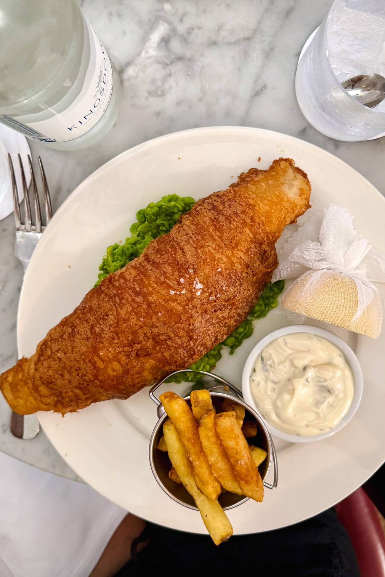 An overhead photo of classic British fish and chips made with battered cod over fresh mushy peas served with a small bucket of fries, half a lemon, and tartar sauce. 