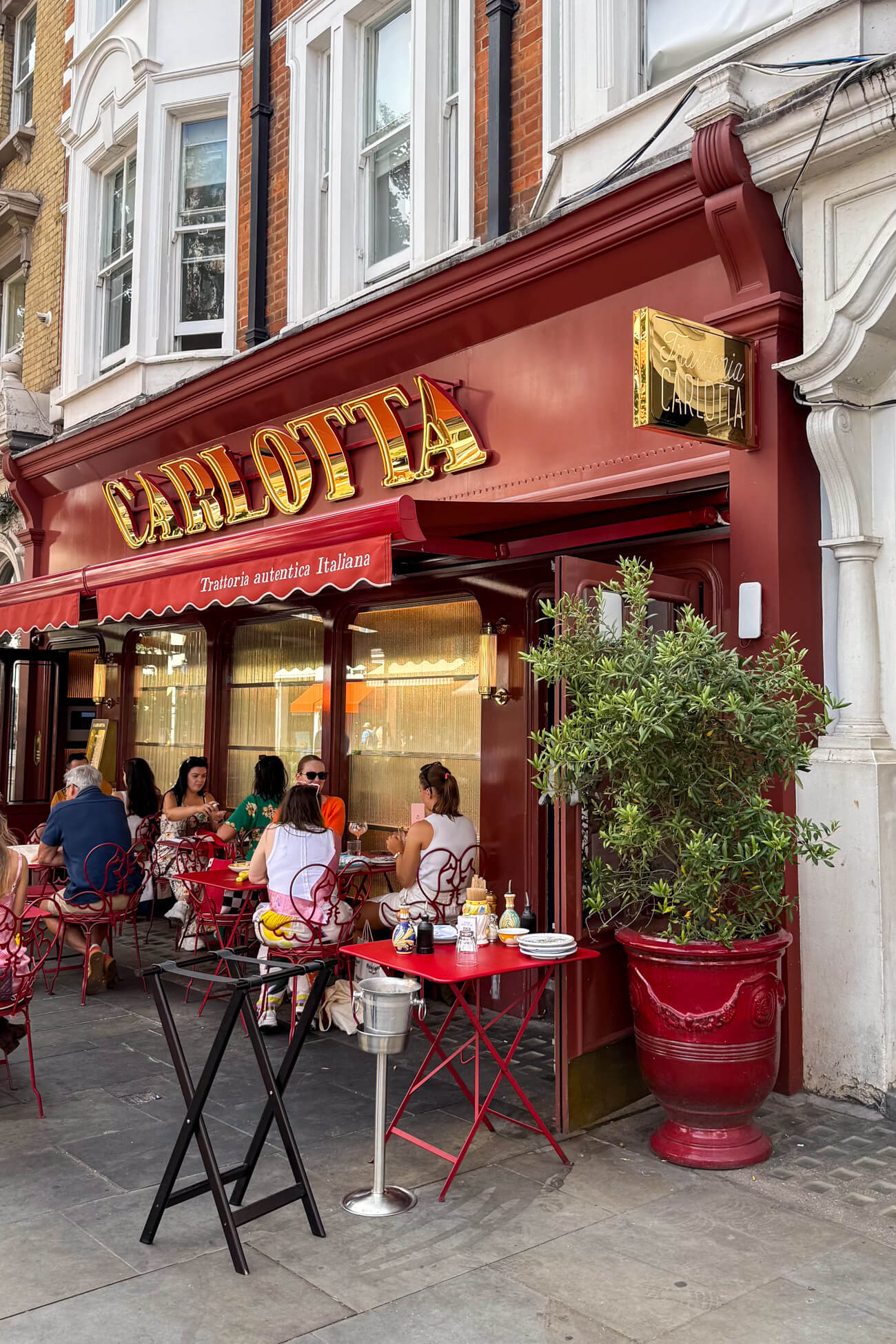 The outside of Carlotta restaurant in Marylebone, London. The restaurant has a red awing with gold letters and people are eating outside. 
