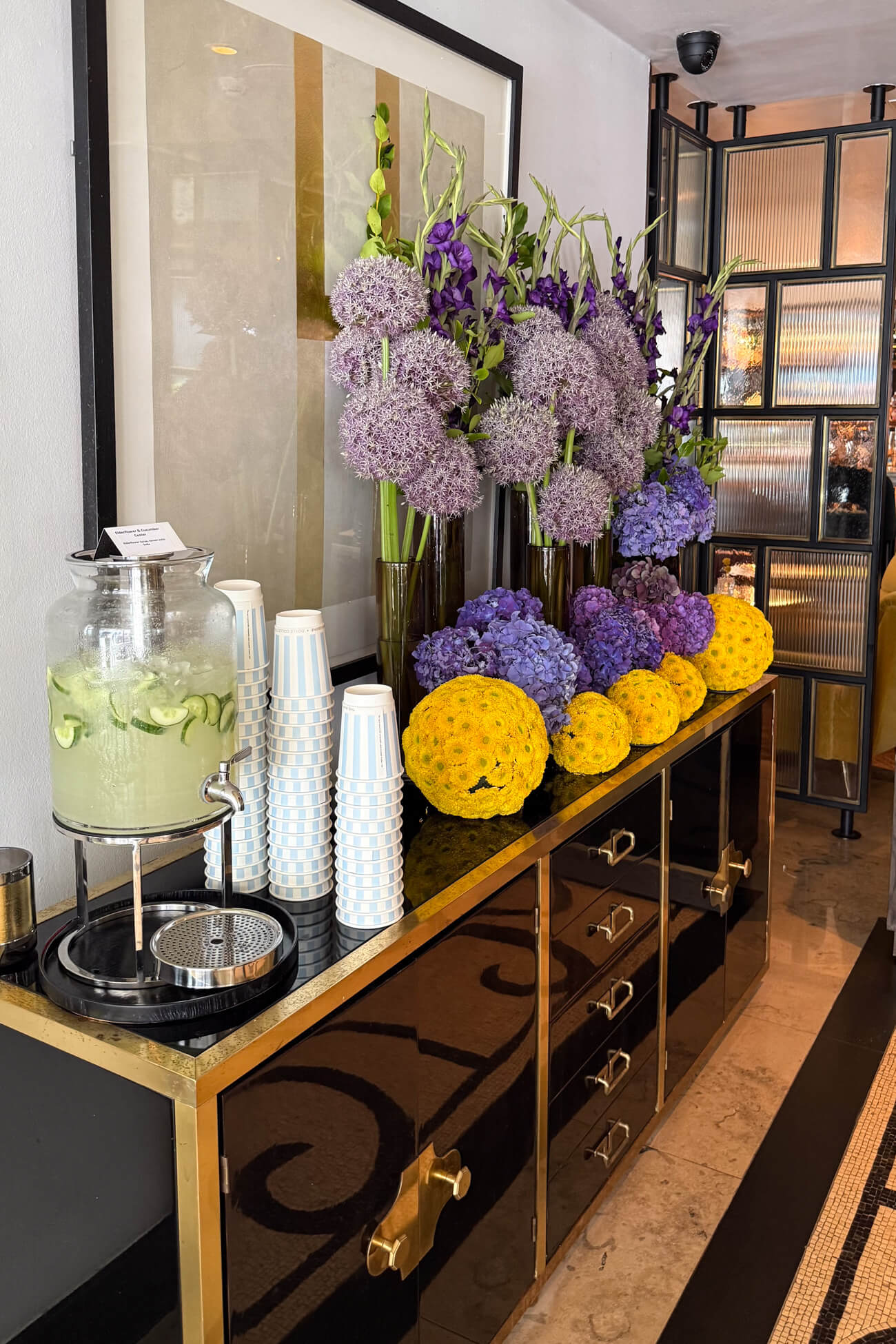 A black and gold buffet table topped with a purple and green flower arrangement and a jug of cucumber beverage with cups in the Marylebone Hotel lobby. 