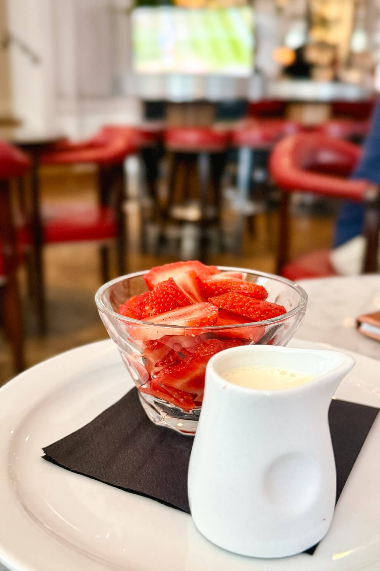 A small glass dish of sliced strawberries with a side of cream four pouring on a restaurant table with tennis on a TV screen in the background. 