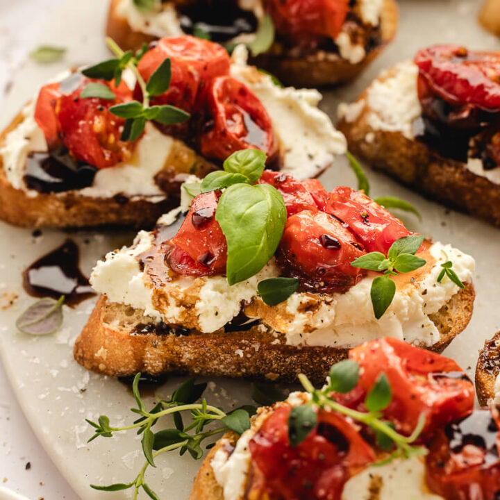 Crostini with whipped ricotta, roasted cherry tomatoes, balsamic drizzle, and basil on a marble serving platter.