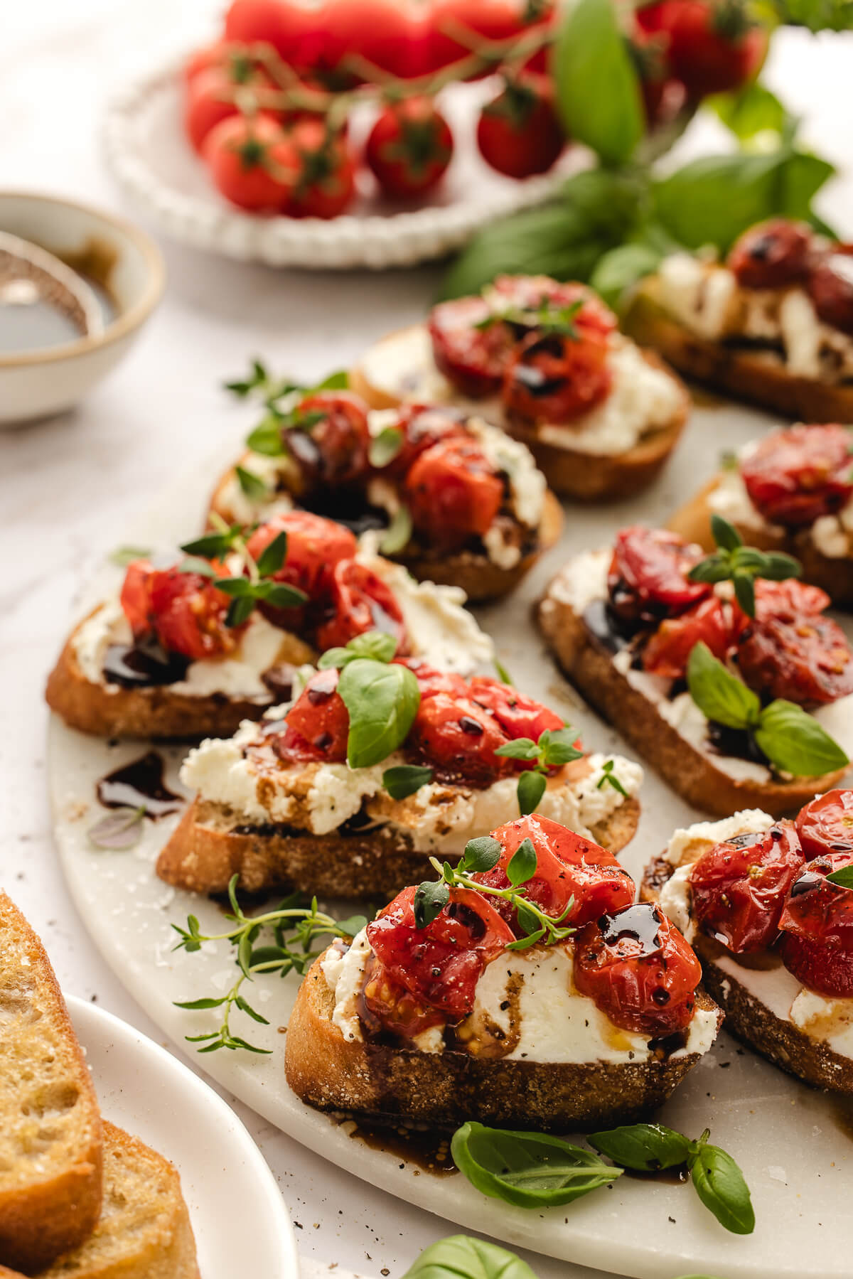 Whipped Ricotta Crostini with Roasted Tomatoes Appetizer Whipped ricotta crostini with roasted cherry tomatoes, balsamic glaze, and fresh herbs arranged on a white serving platter.