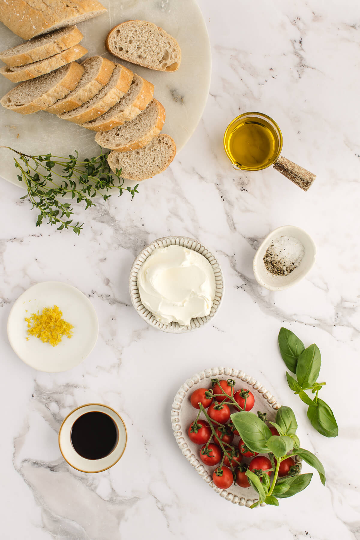 crostini flat lay An overhead photo of the ingredients for whipped ricotta crostini with cherry tomatoes. Clockwise from the top: sliced baguette, olive oil, salt and pepper, basil, cherry tomatoes, balsamic, ricotta, and fresh thyme.