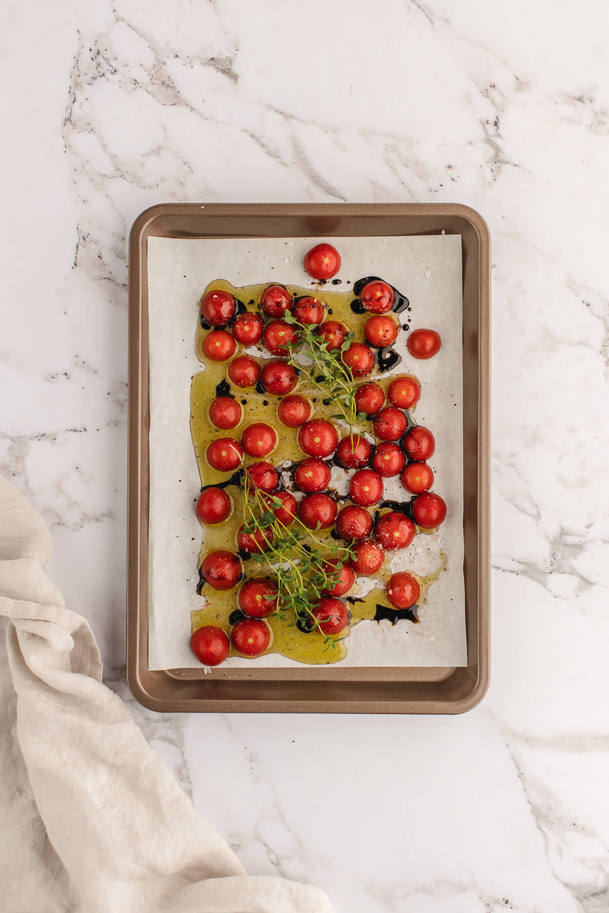 An overhead photo of cherry tomatoes with olive oil and herbs on a parchment-lined sheet pan.