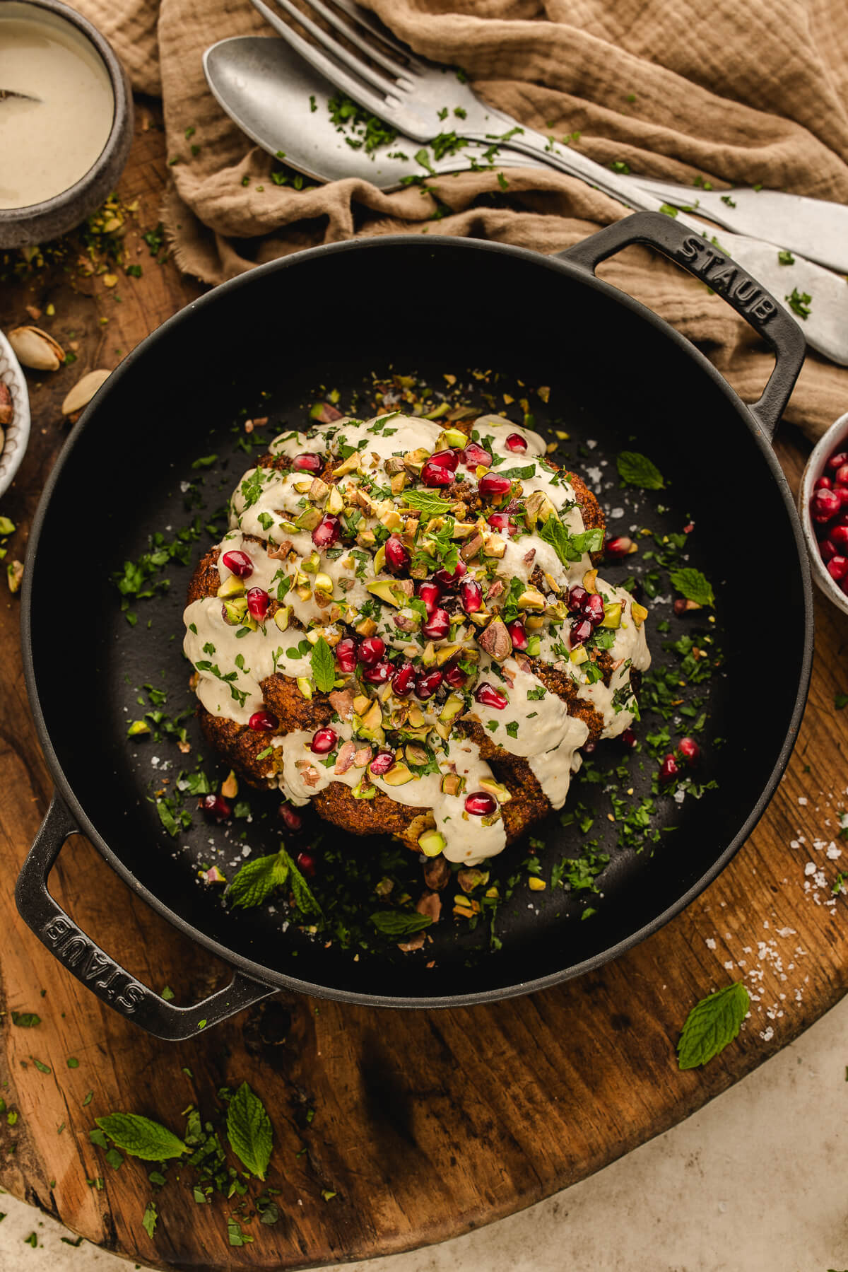 Whole roasted cauliflower served in a Staub cast-iron skillet topped with creamy tahini sauce, chopped pistachios, pomegranate seeds, and fresh herbs.