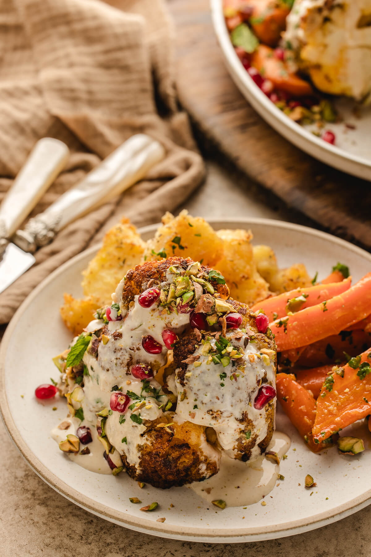 Plated serving of whole roasted cauliflower with tahini sauce, pistachios, pomegranate seeds, and roasted carrots — an elegant vegetarian holiday meal.