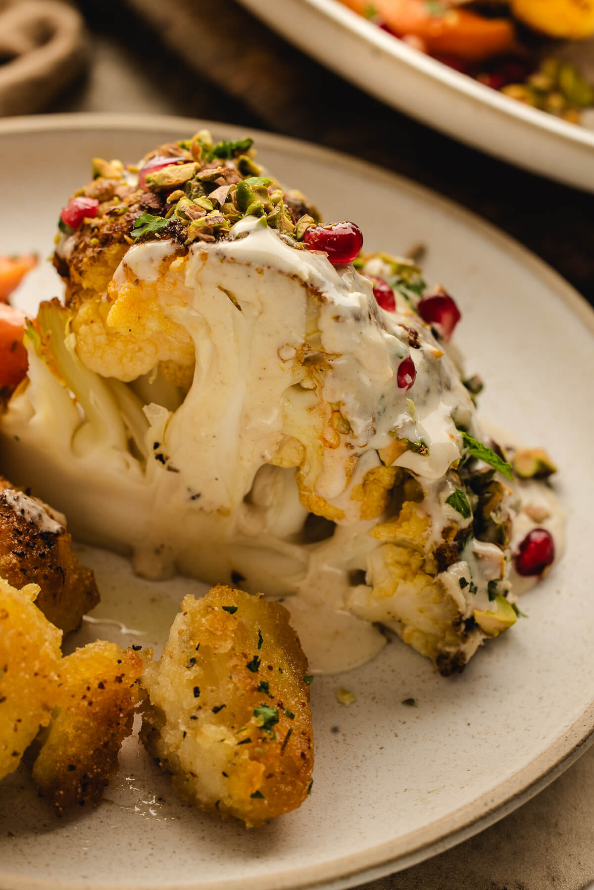 Close-up of a slice of roasted cauliflower topped with creamy tahini sauce, chopped pistachios, and pomegranate seeds, served with crispy potatoes.