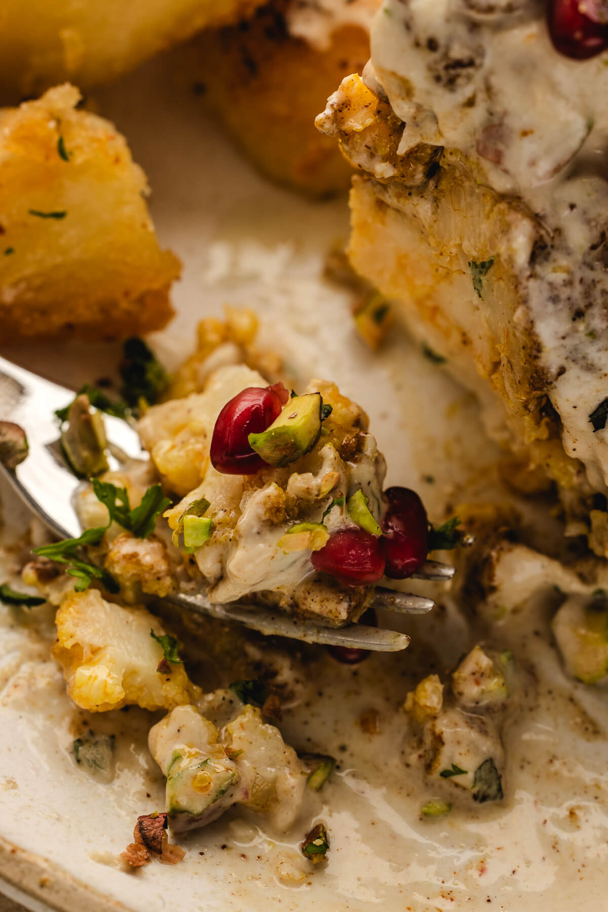 Close-up bite of roasted cauliflower with creamy tahini sauce, pistachios, and pomegranate seeds on a fork.