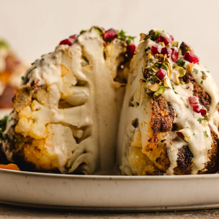 A side view of a whole roasted cauliflower with a slice out so that you can see inside. The cauliflower is dressed with tahini dressing, pomegranate arils and pistachios, making it a festive vegetarian main course for holidays.