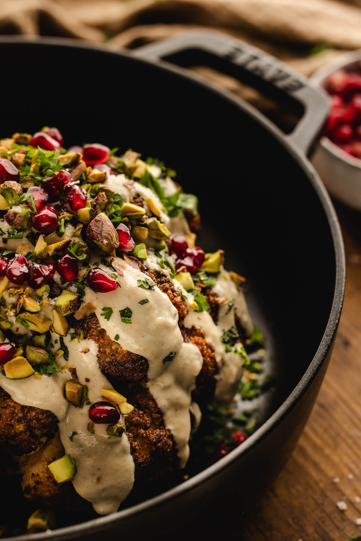 Whole roasted cauliflower in a cast iron skillet, topped with creamy tahini sauce, chopped pistachios, pomegranate seeds, and fresh herbs.