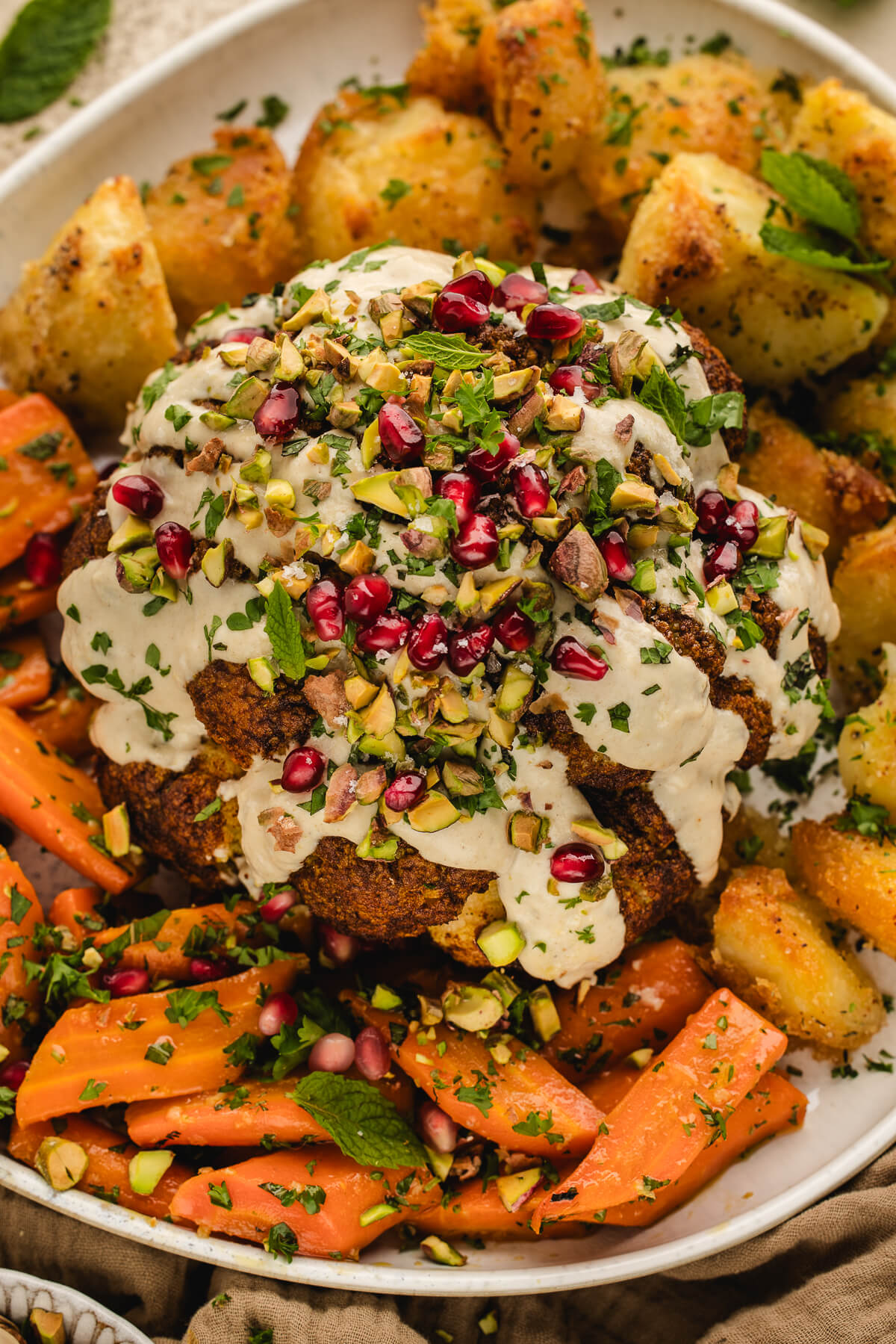 Whole roasted cauliflower with tahini sauce, pistachios, and pomegranate seeds served on a platter with roasted carrots and potatoes — a beautiful vegetarian holiday centerpiece.