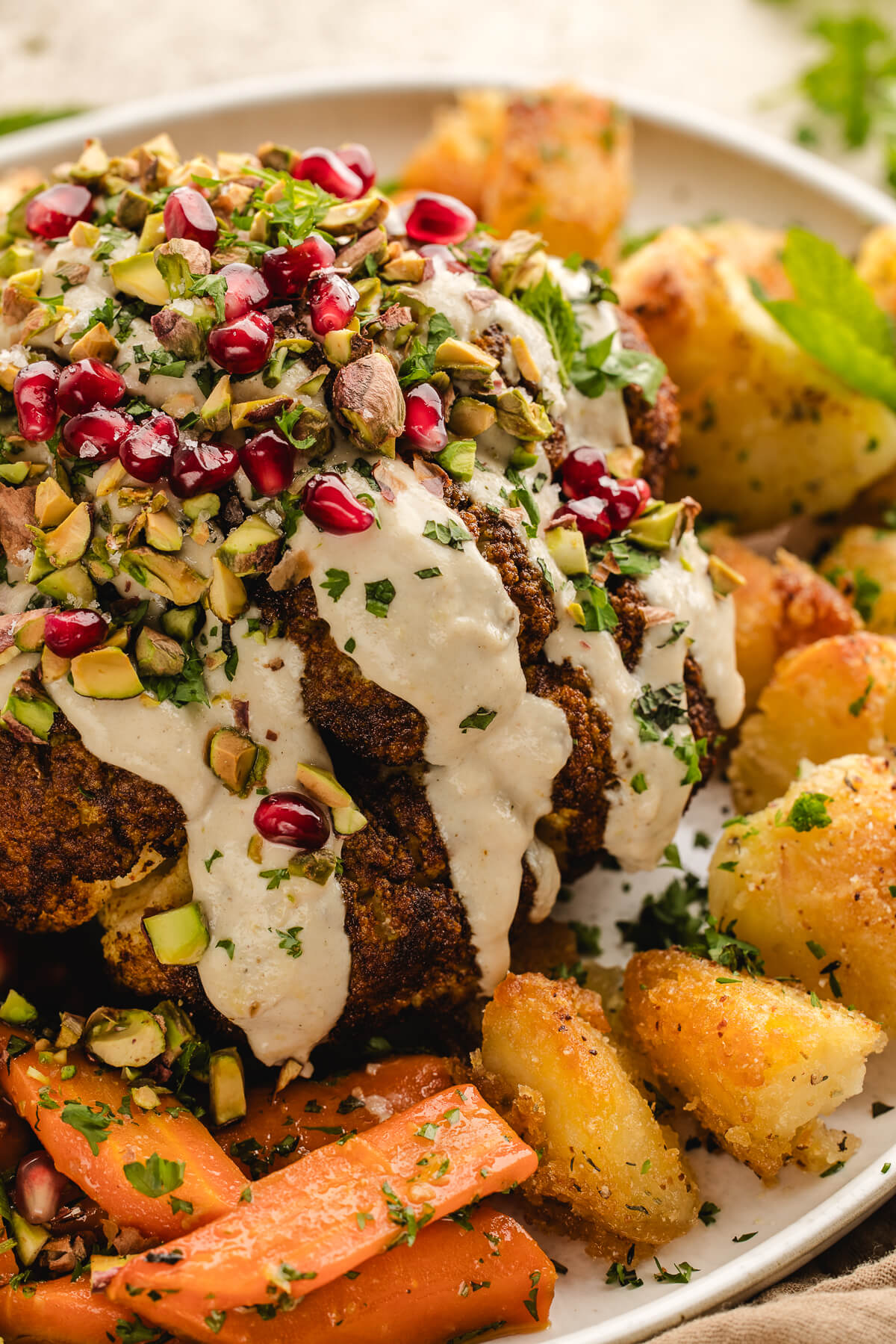 A whole roasted cauliflower topped with creamy tahini dressing, pomegranate, pistachios and herbs on a platter with roasted potatoes and carrots.