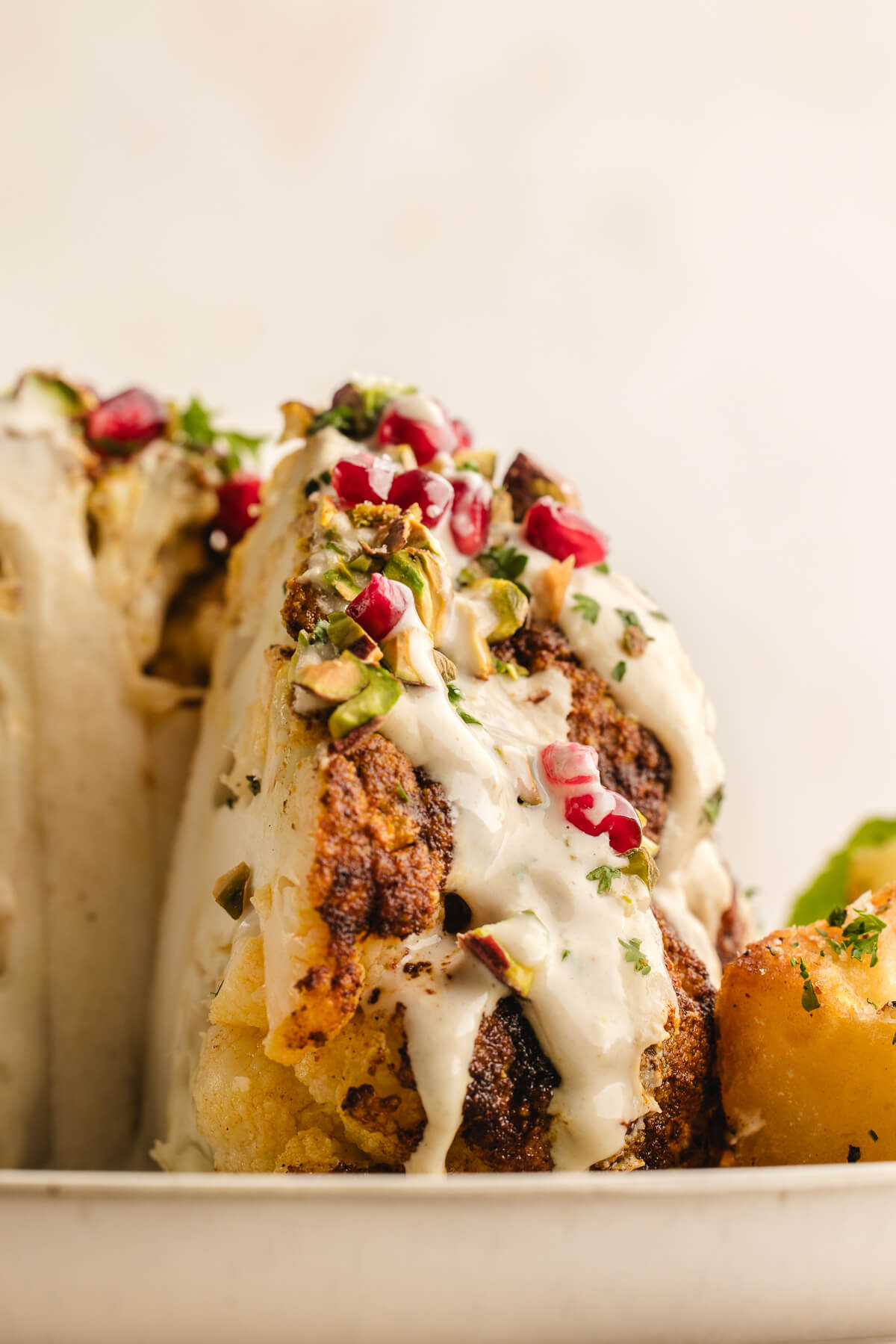 Close-up of roasted cauliflower wedge drizzled with tahini sauce and topped with pomegranate seeds, chopped pistachios, and herbs.