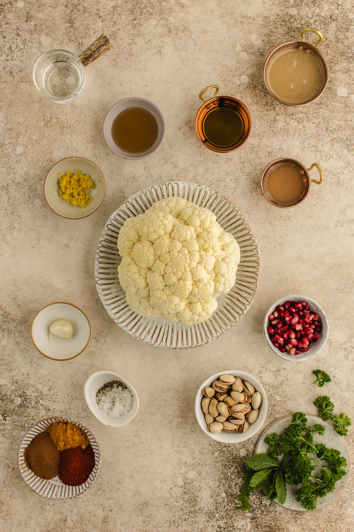 ngredients for whole roasted cauliflower including fresh cauliflower, tahini, olive oil, lemon zest, garlic, herbs, pistachios, and pomegranate seeds arranged on a neutral surface.